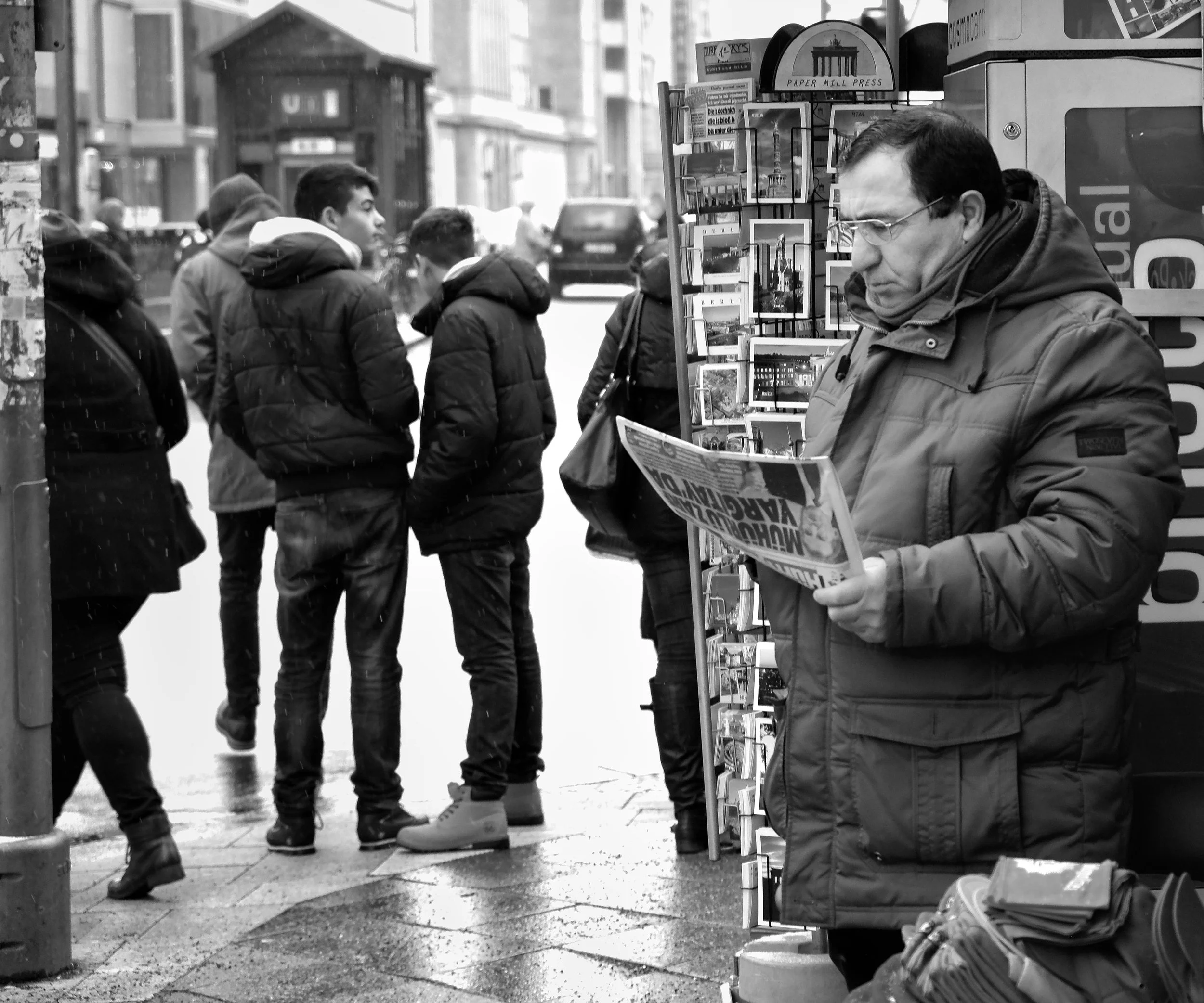 Black and white street photograph of a man reading a newspaper on a rainy street in Berlin