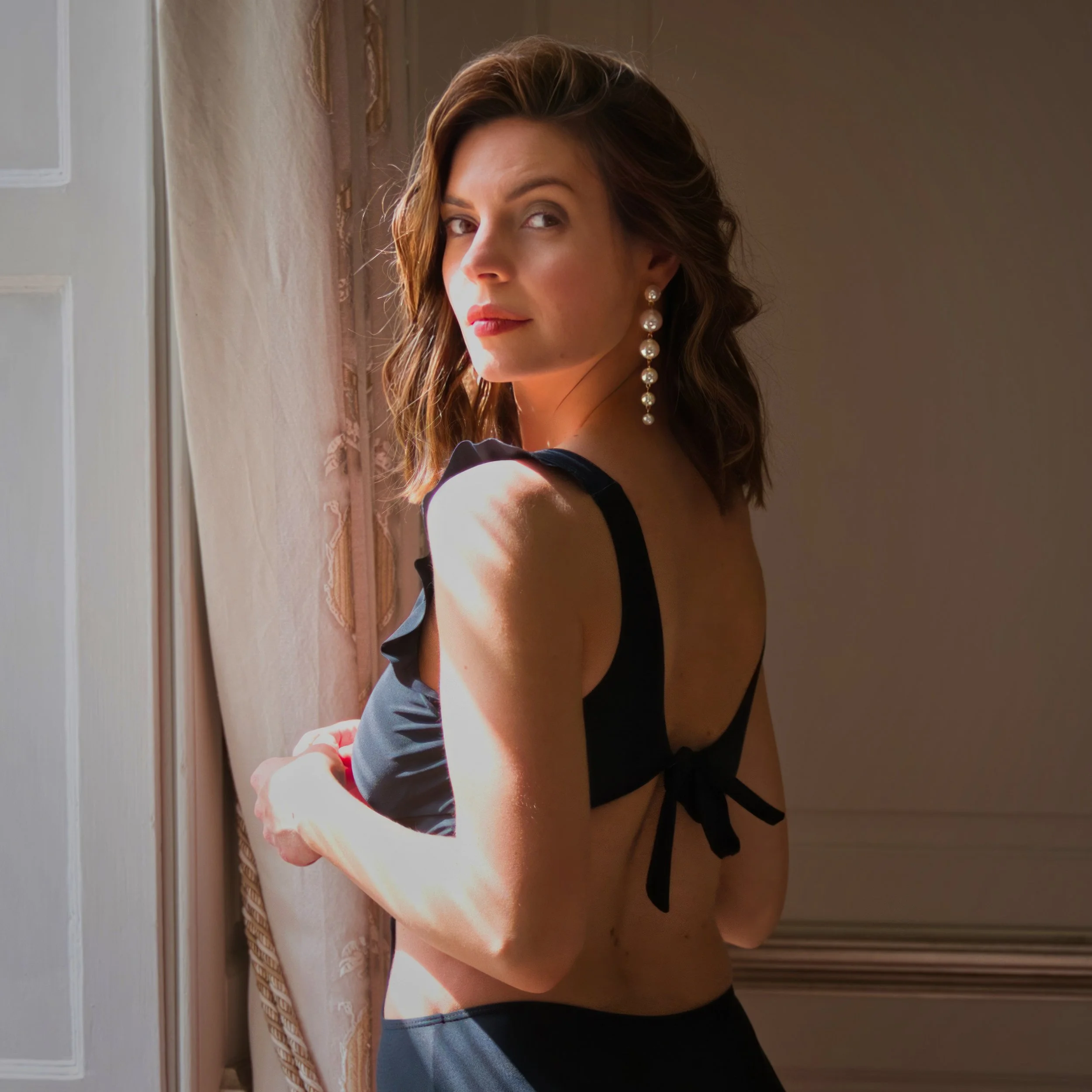 A woman in a black swimsuit and pearl drop earrings looks over her shoulder beside a window, illuminated by warm natural light.