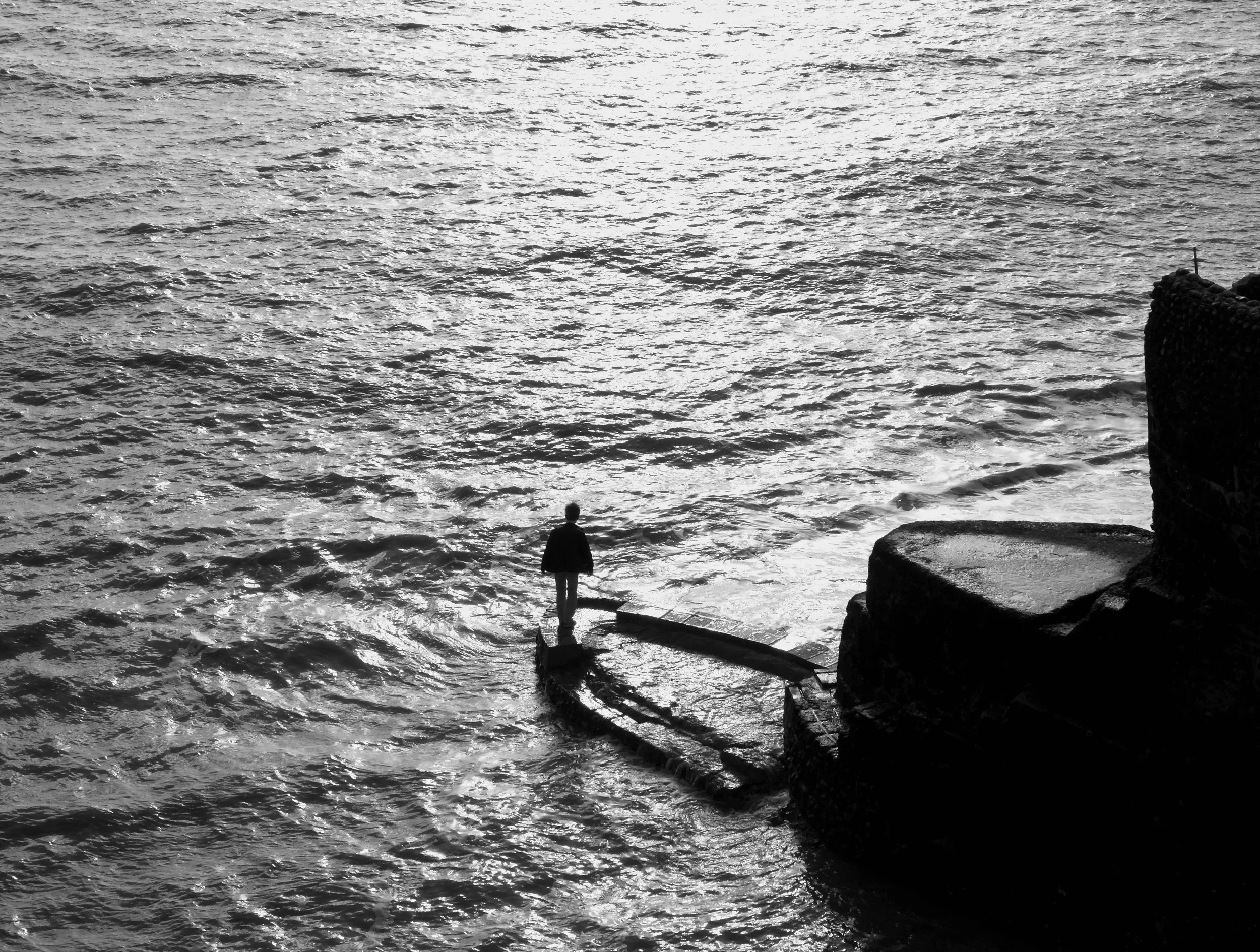 Small solitary figure standing at the edge of a flooded stone platform beneath dark cliffs, surrounded by shimmering sea water in Brighton
