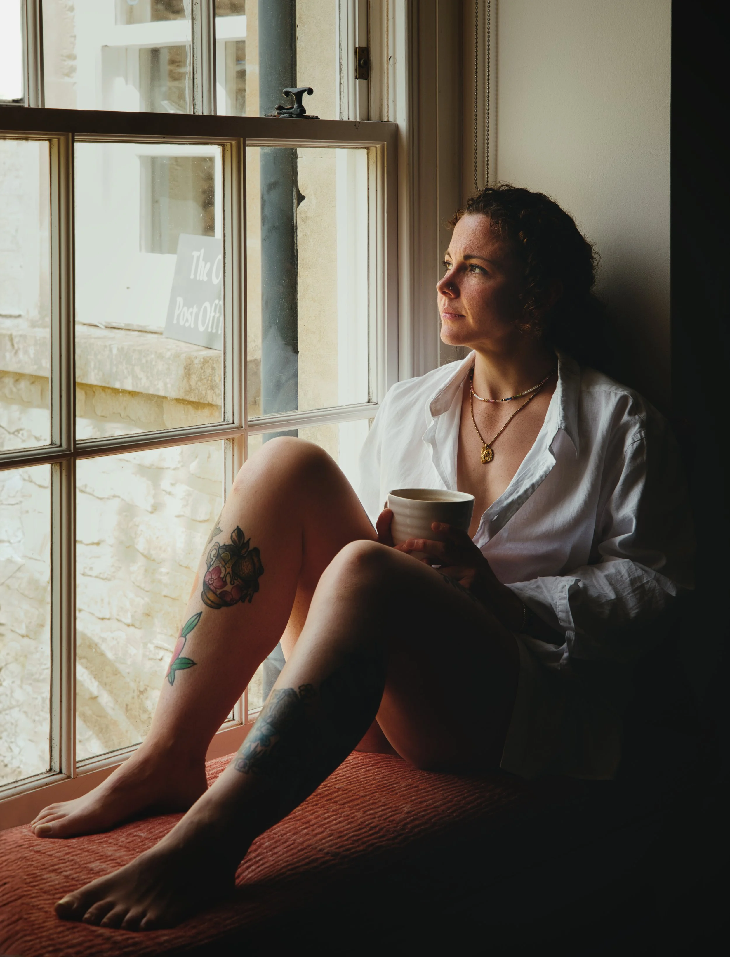 Woman sitting on a window seat holding a coffee cup, photographed in natural light during a portrait session in Gloucestershire.