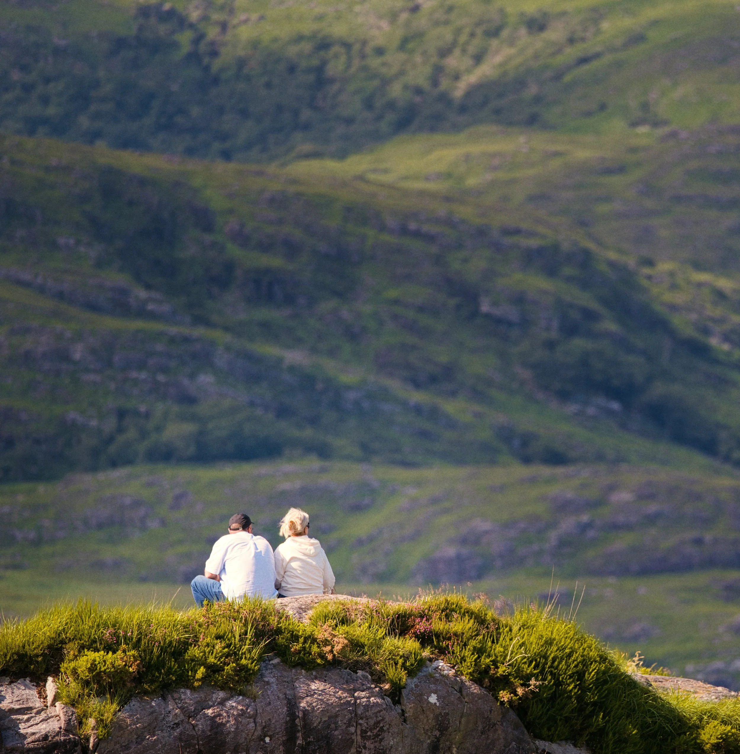 Couple sitting side by side on a rocky outcrop with the mountains of rural Ireland in the background.