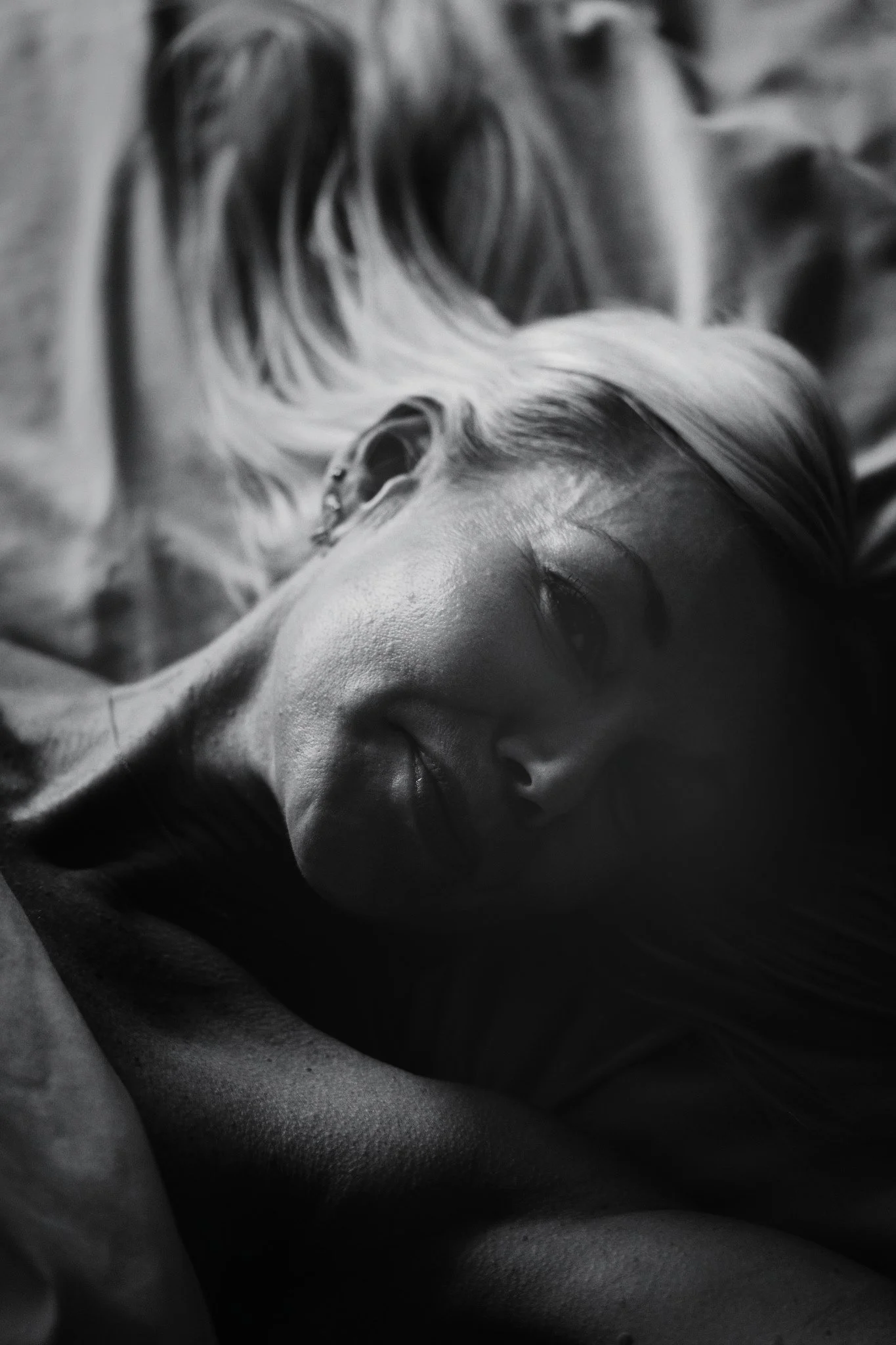 Intimate black and white close-up portrait of a blonde woman reclining on a bed, with soft light illuminating her face against deep, atmospheric shadows.