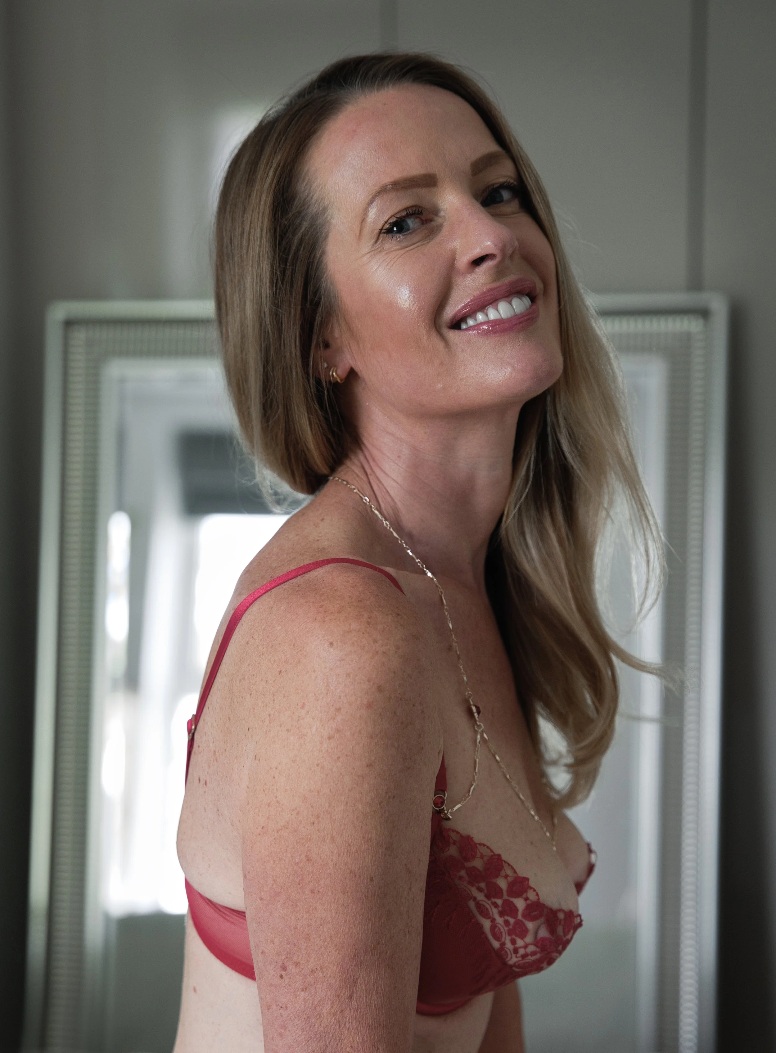 A woman with long, wavy, light brown hair smiling at the camera, wearing a red lingerie top with lace details and a thin necklace, standing indoors near a mirror with a gray wall in the background.