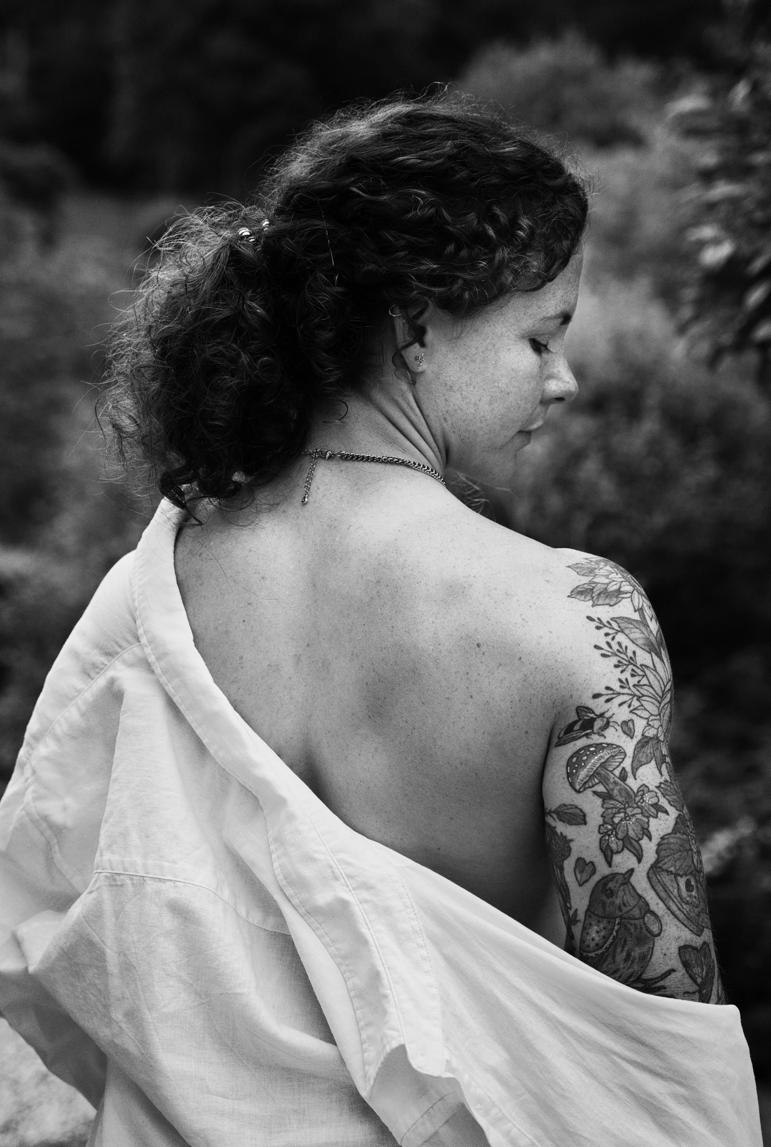 Black and white portrait of a woman with tattoos looking over her shoulder while an oversized white shirt falls from her back.