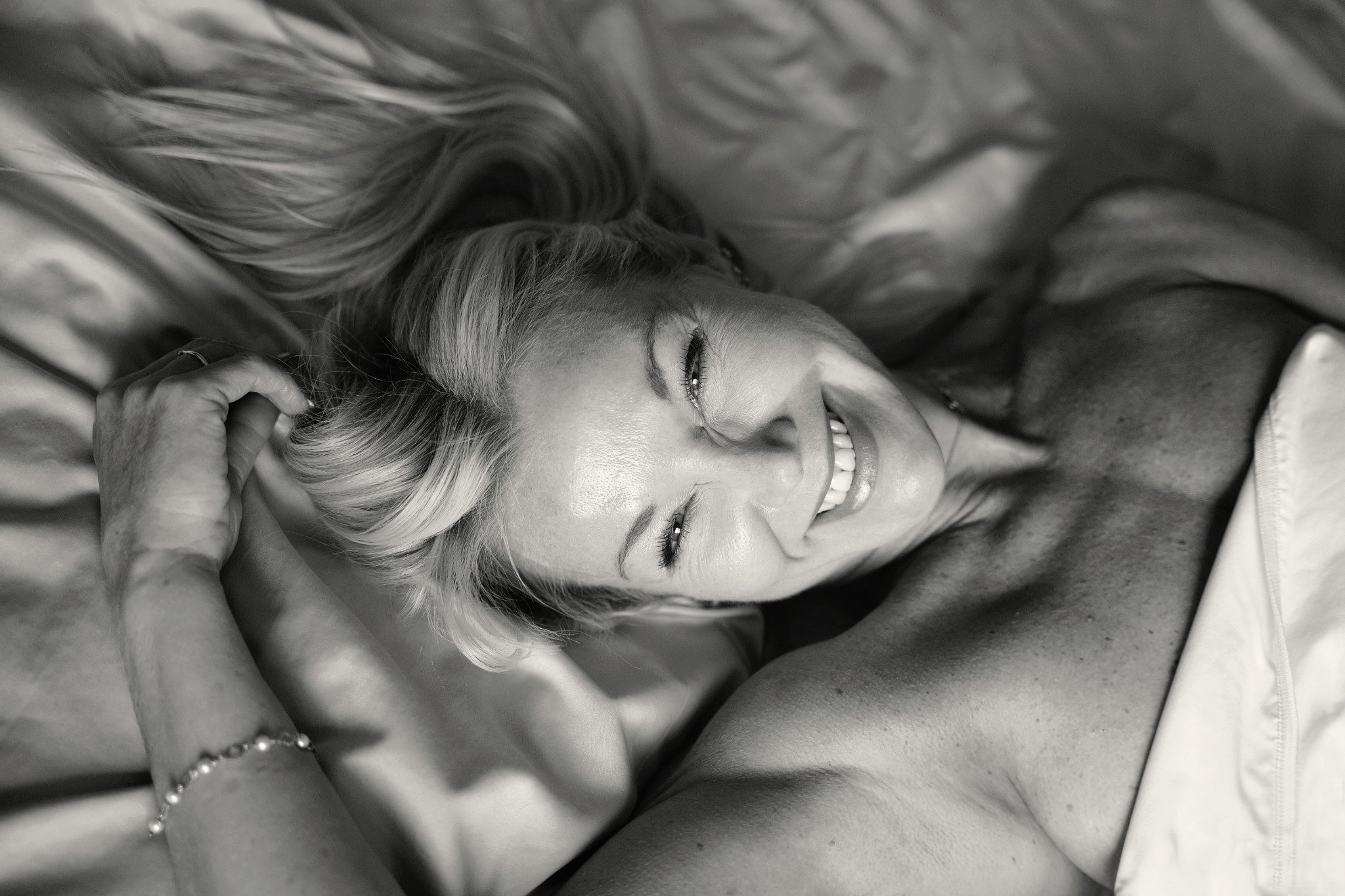 Black and white boudoir portrait of a smiling blonde woman lying on a bed, with her hair spread across the pillows and her hands resting above her head.