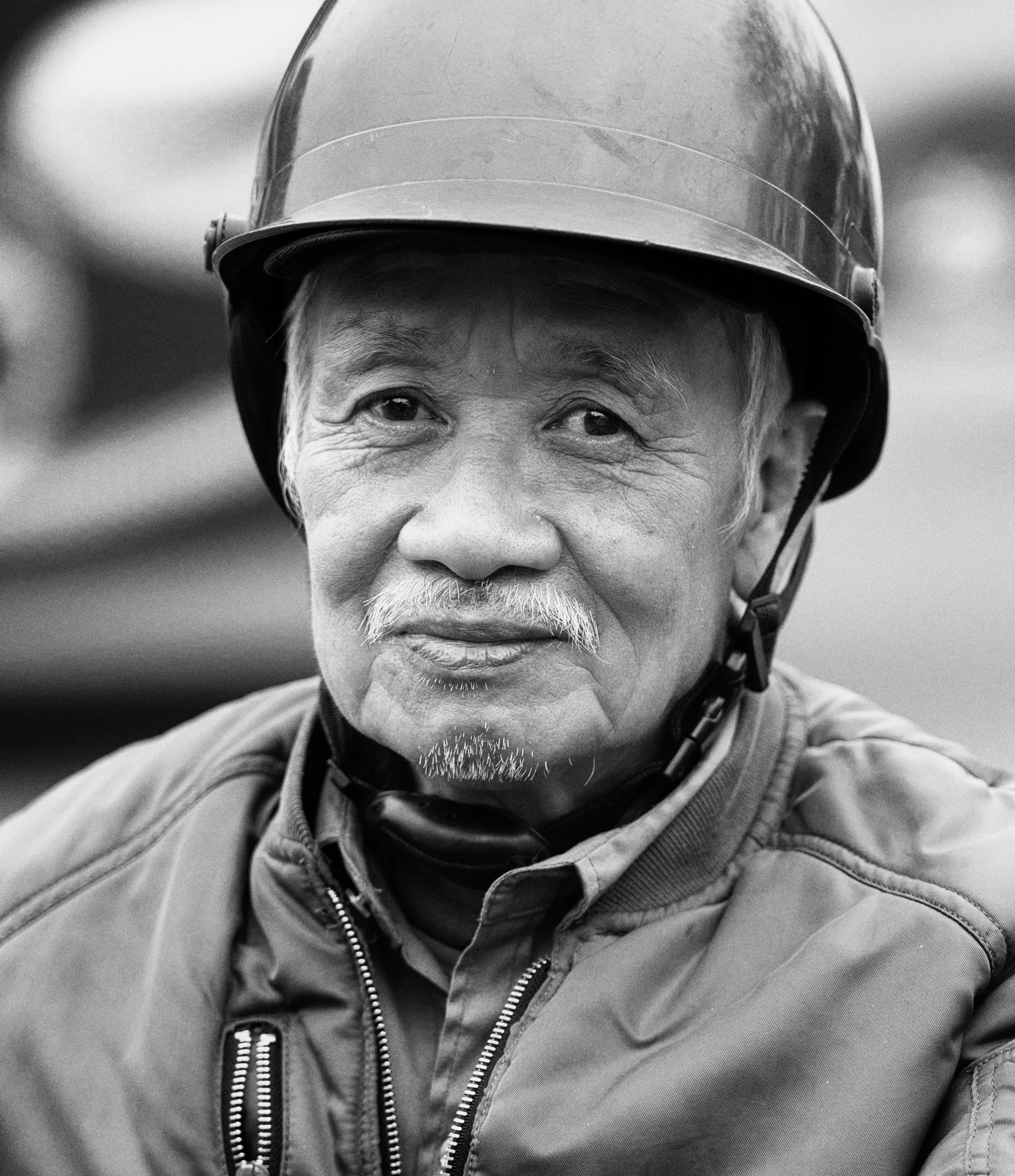 Black and white portrait of an older man in Hanoi wearing a helmet with calm expression and direct gaze