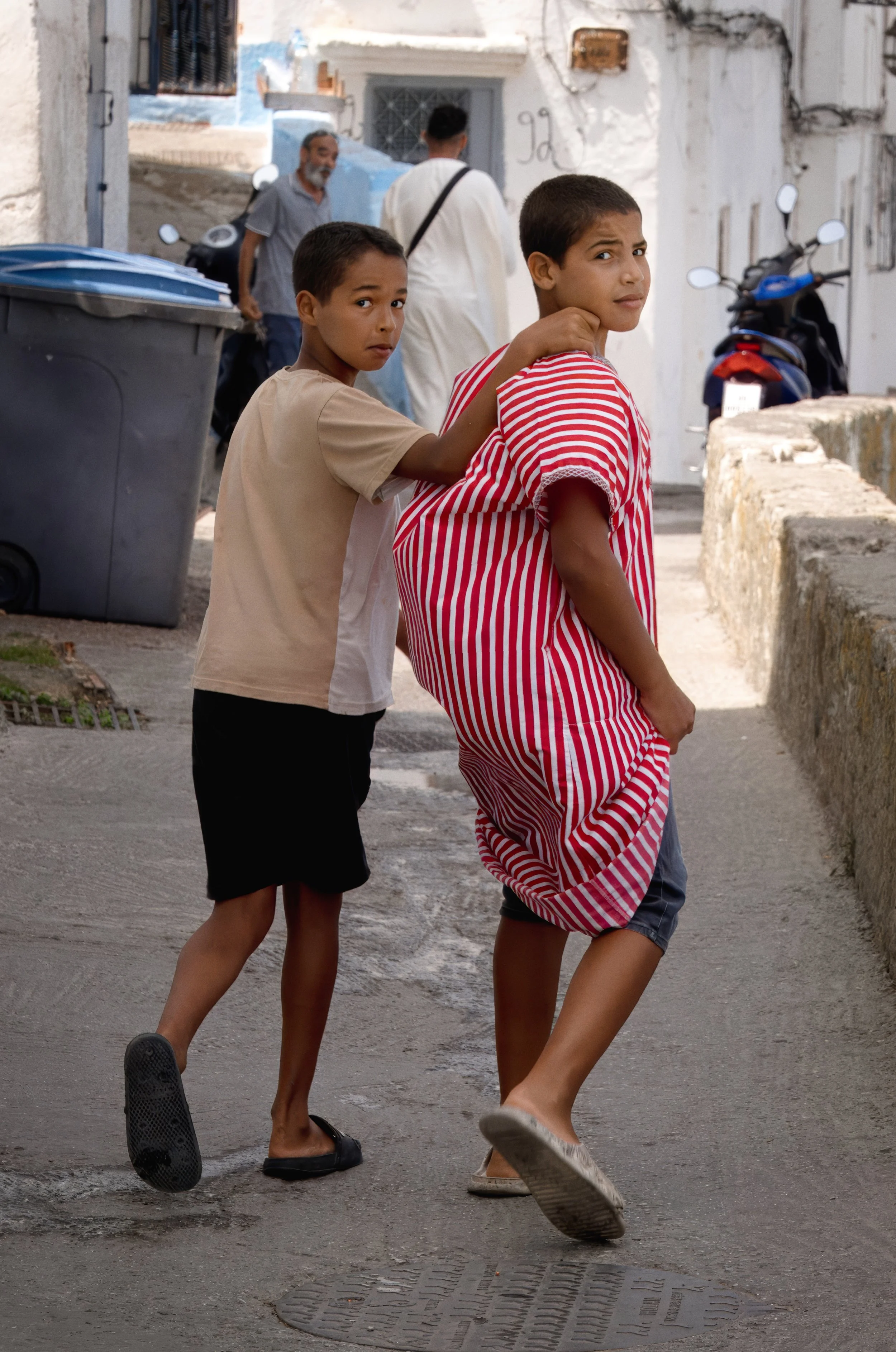 Two boys walking through a narrow street in Tangier, one carrying a striped sack over his shoulder while both look back toward the camera