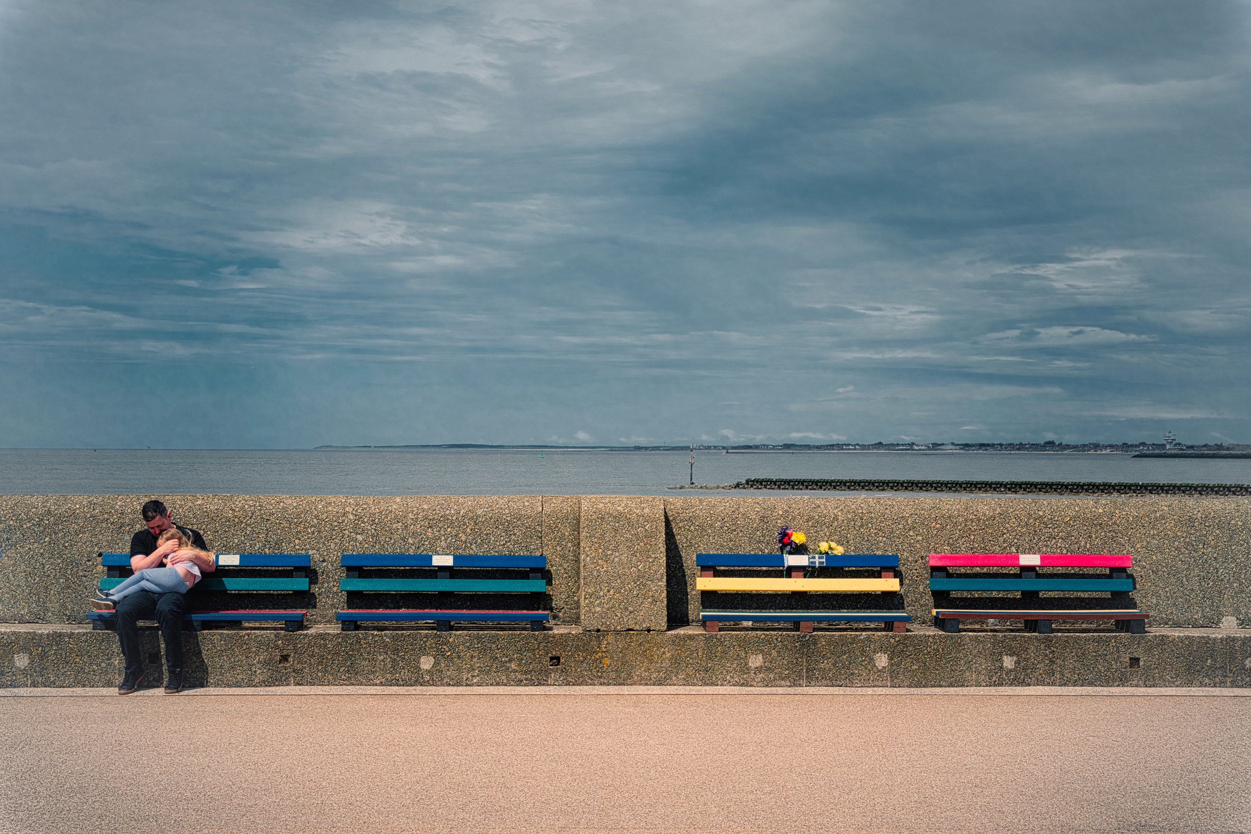 A father and daughter share a quiet moment on a seafront bench, framed by empty benches, flowers and an expansive coastal sky.
