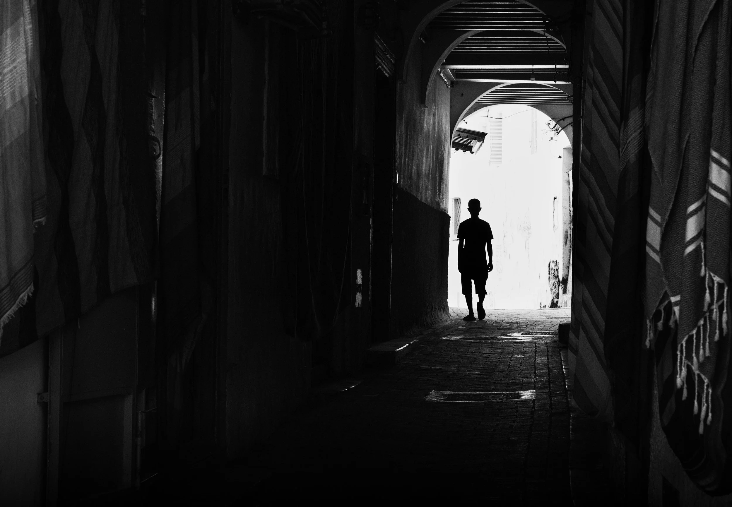 Black and white silhouette of a person walking through a narrow passageway toward bright light