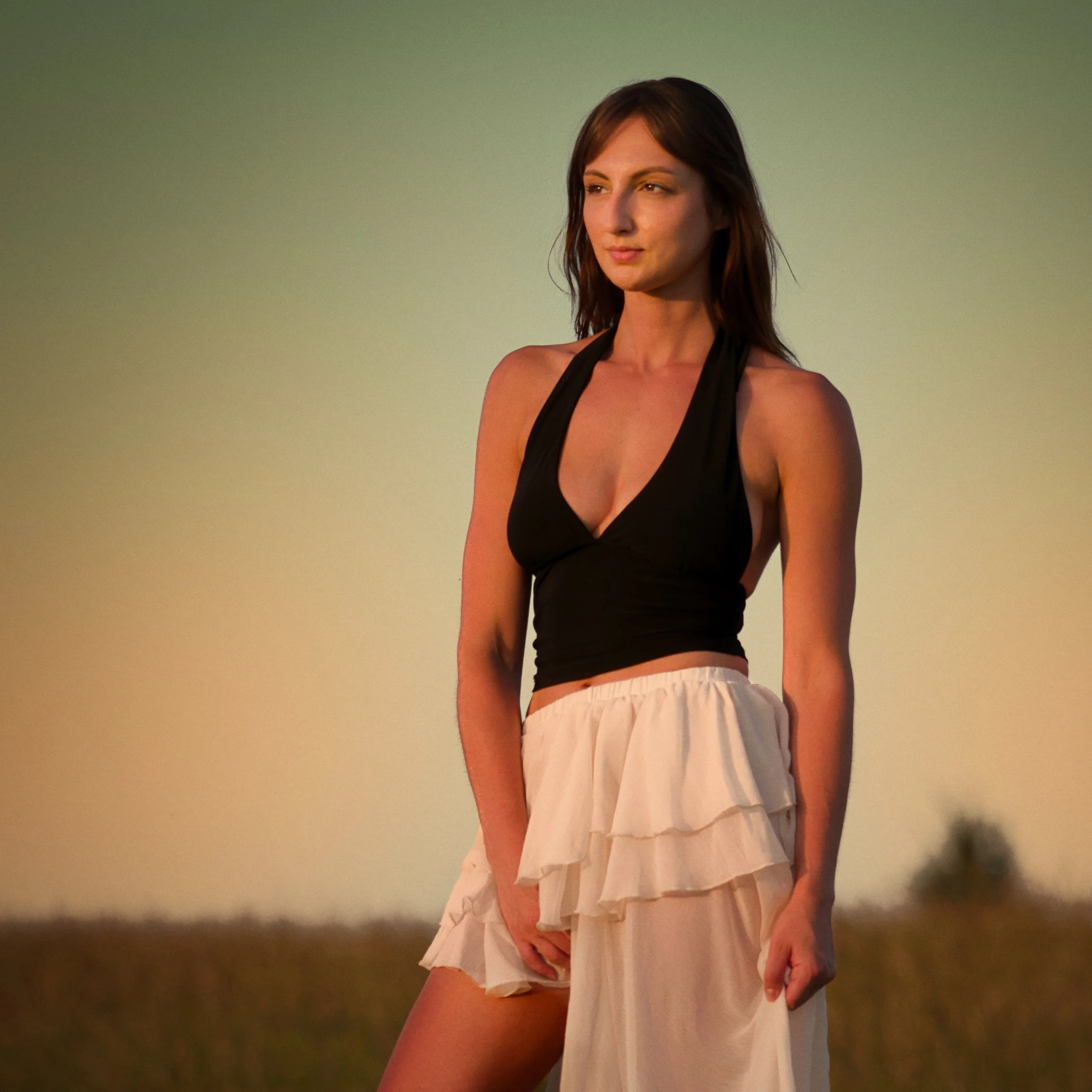 Outdoor portrait of a woman in a black halter top and flowing white skirt standing in a meadow during golden hour, photographed in warm natural light.