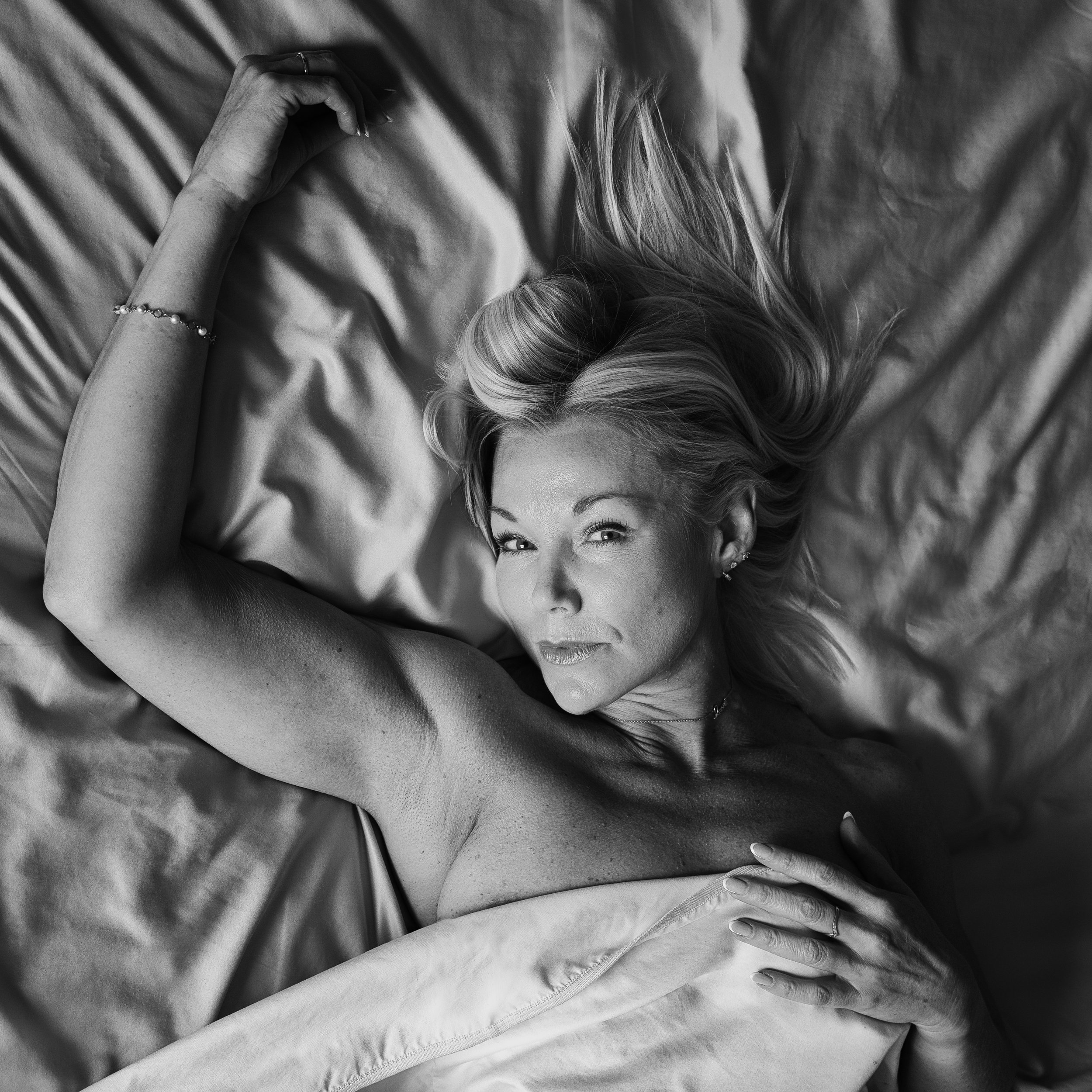 Intimate black and white boudoir portrait of a blonde woman lying in bed, looking into the camera while wrapped in a sheet.