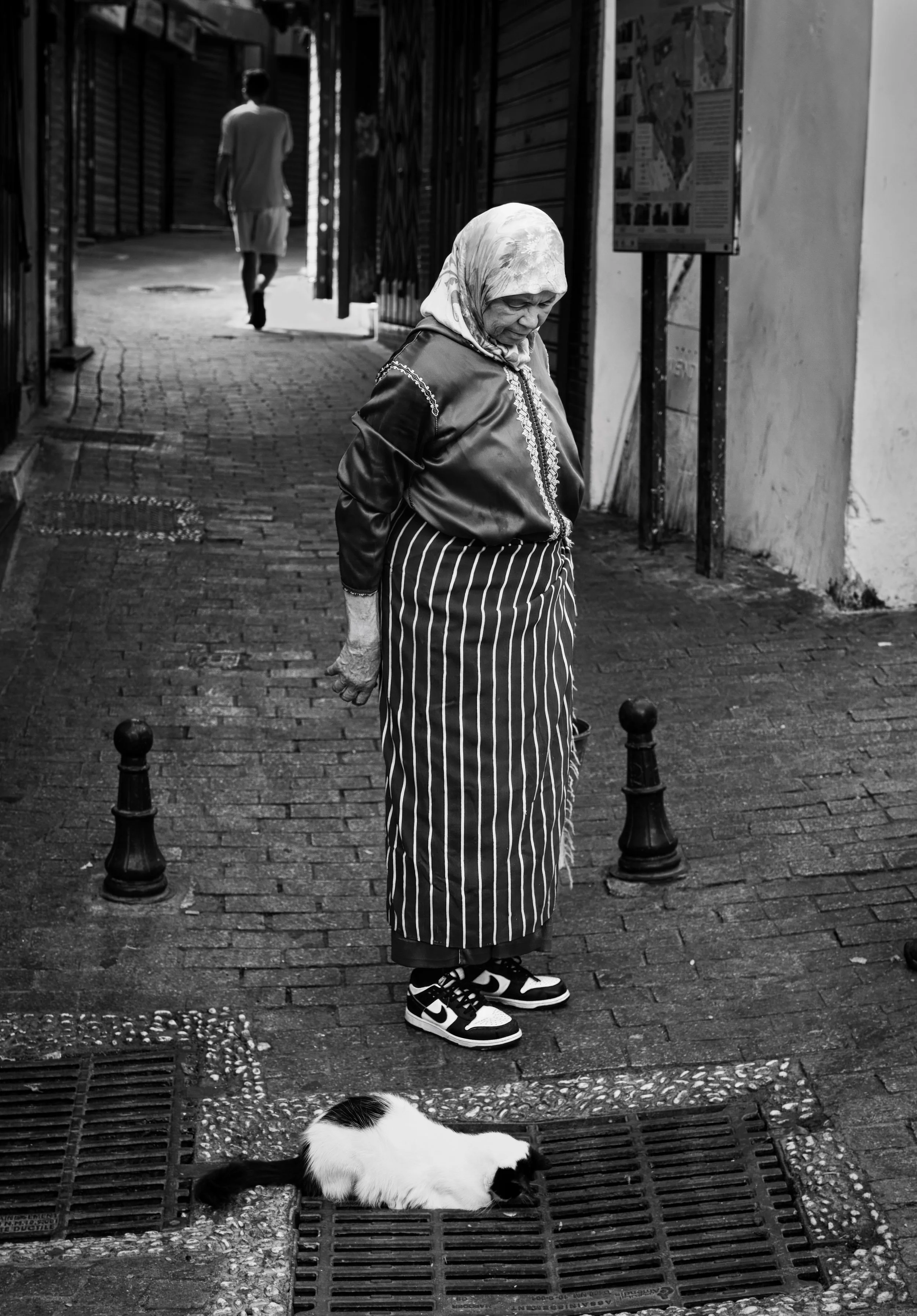 Elderly Moroccan woman in Nike trainers looking at a resting cat in a narrow market alley