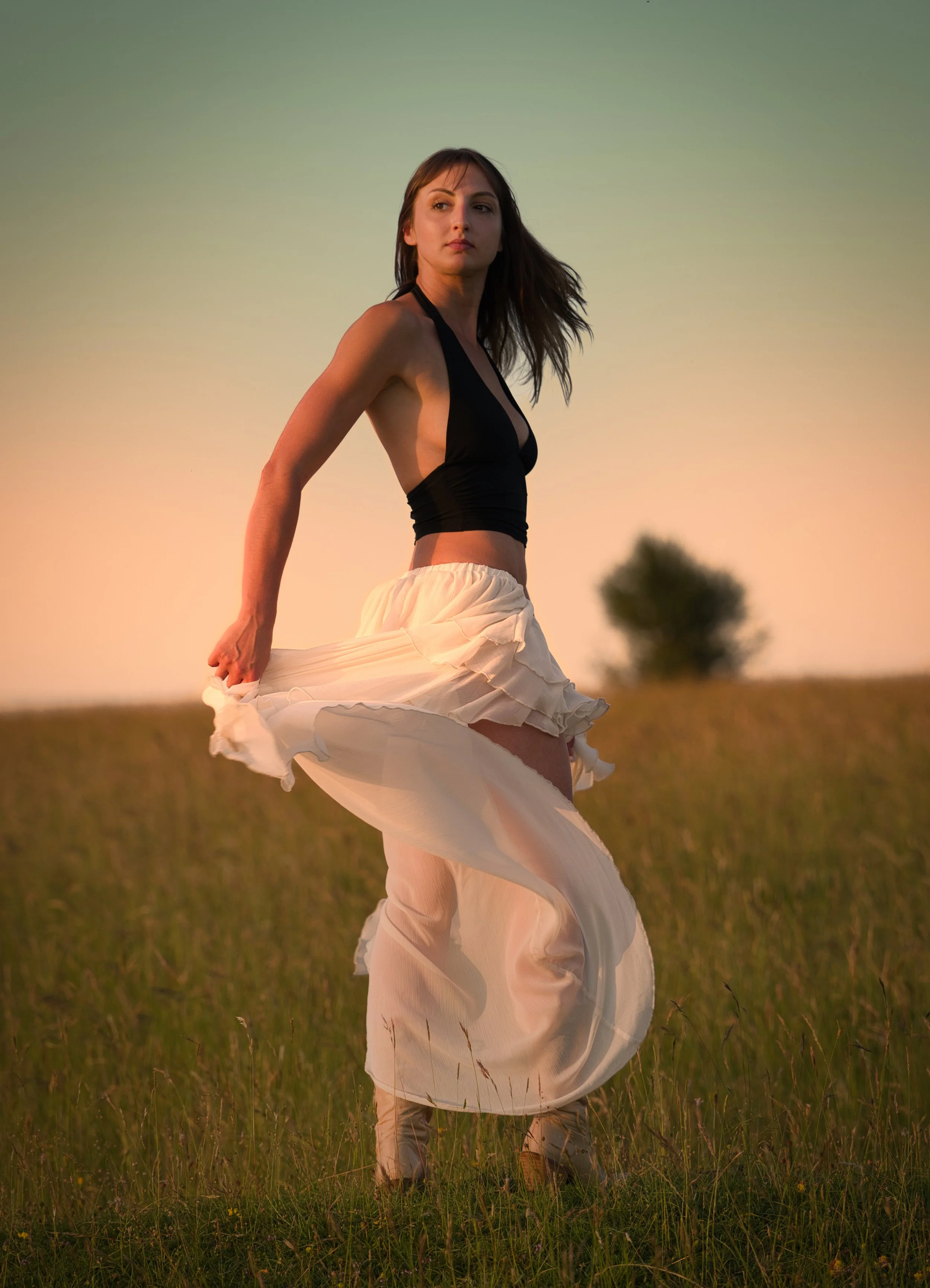 Elegant outdoor portrait of a woman holding a flowing white skirt in a meadow at golden hour, photographed in warm natural light.