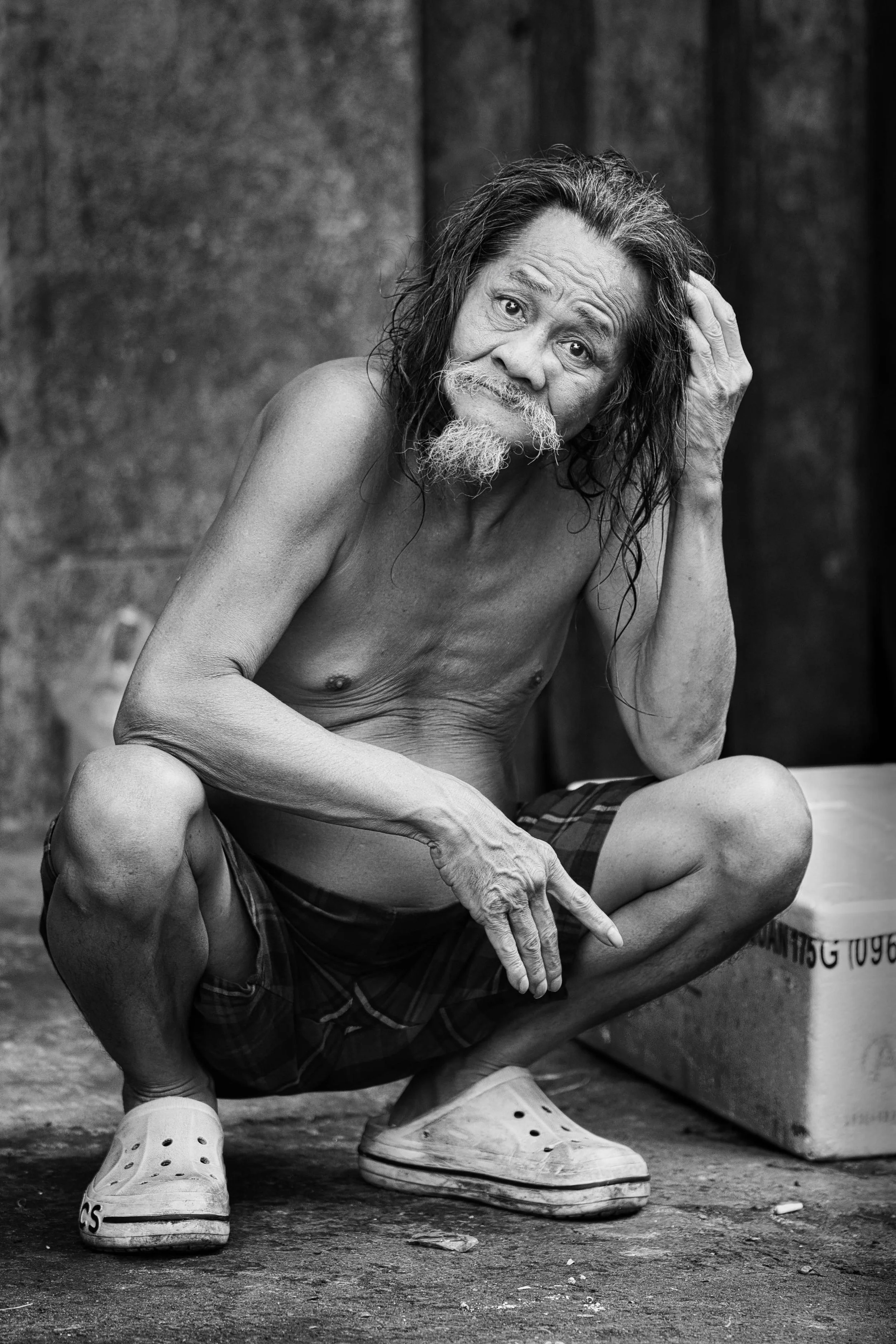 Bare-chested older man with long hair and a pointed white beard crouching on a street in Ho Chi Minh City, looking directly toward the camera