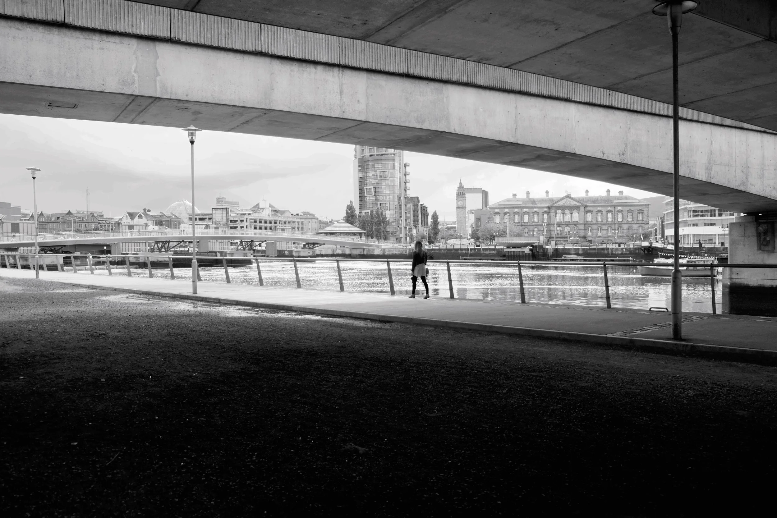 Black and white photograph of a lone figure walking beside the river beneath a bridge in Belfast