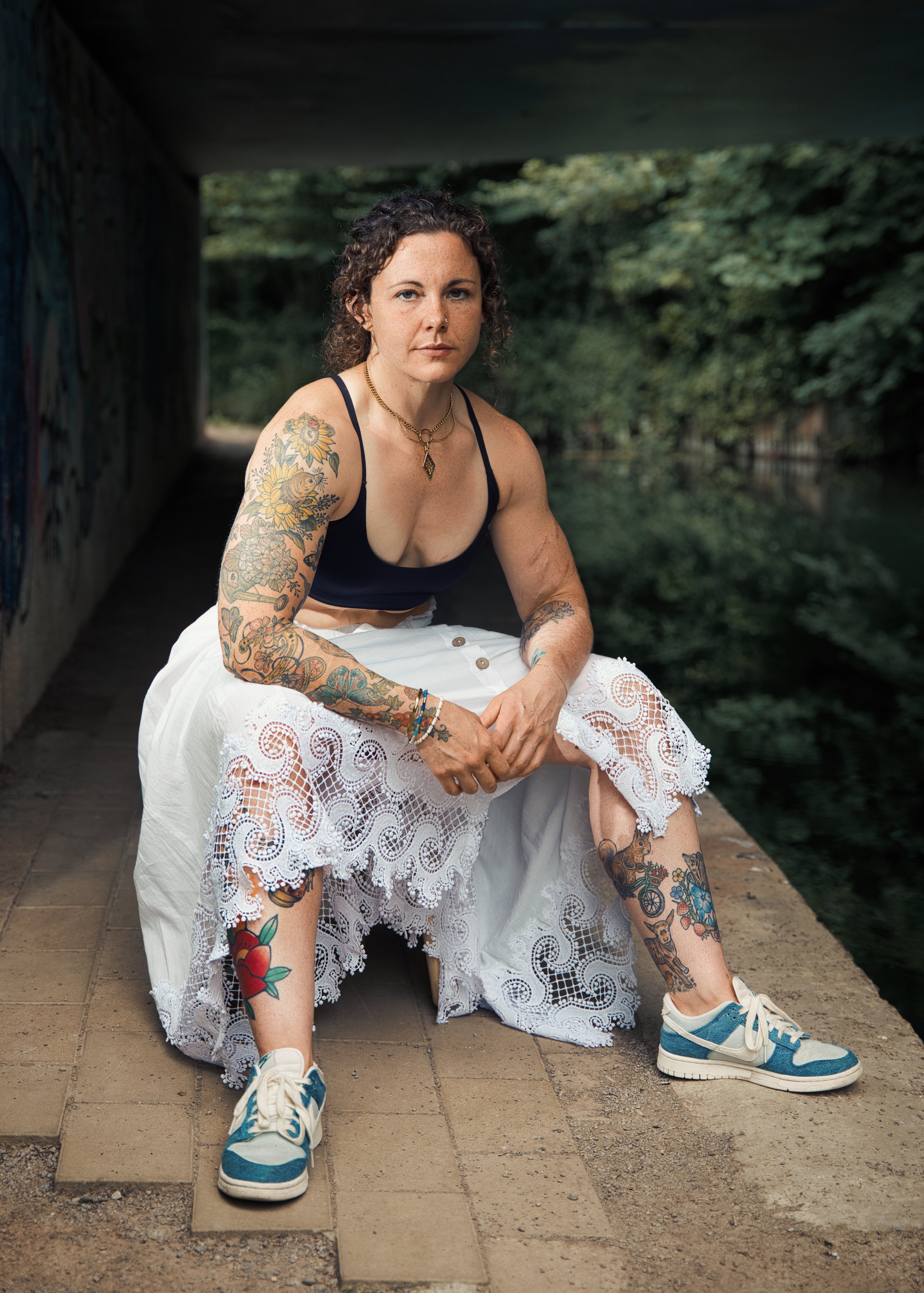 Portrait of a tattooed woman sitting beneath a canal underpass wearing a lace skirt and trainers, photographed in natural light.