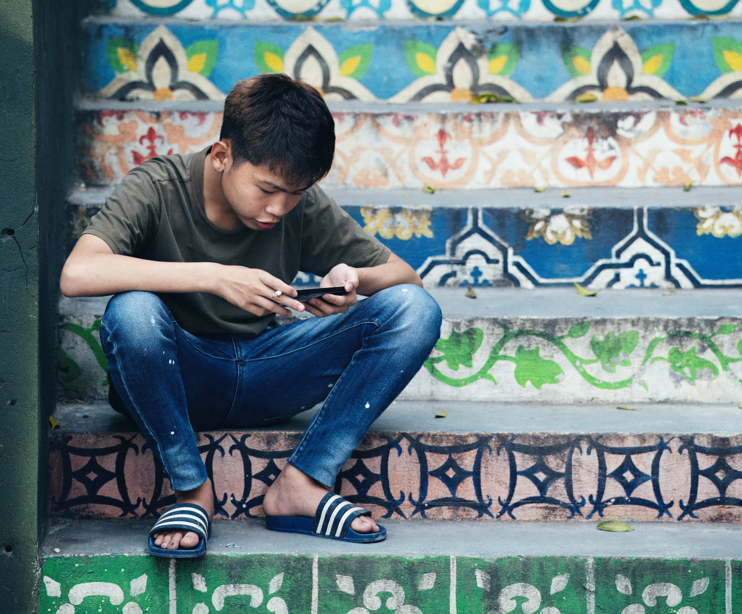 Young man sitting on patterned tiled steps in Ninh Binh, focused on his phone with a cigarette in hand