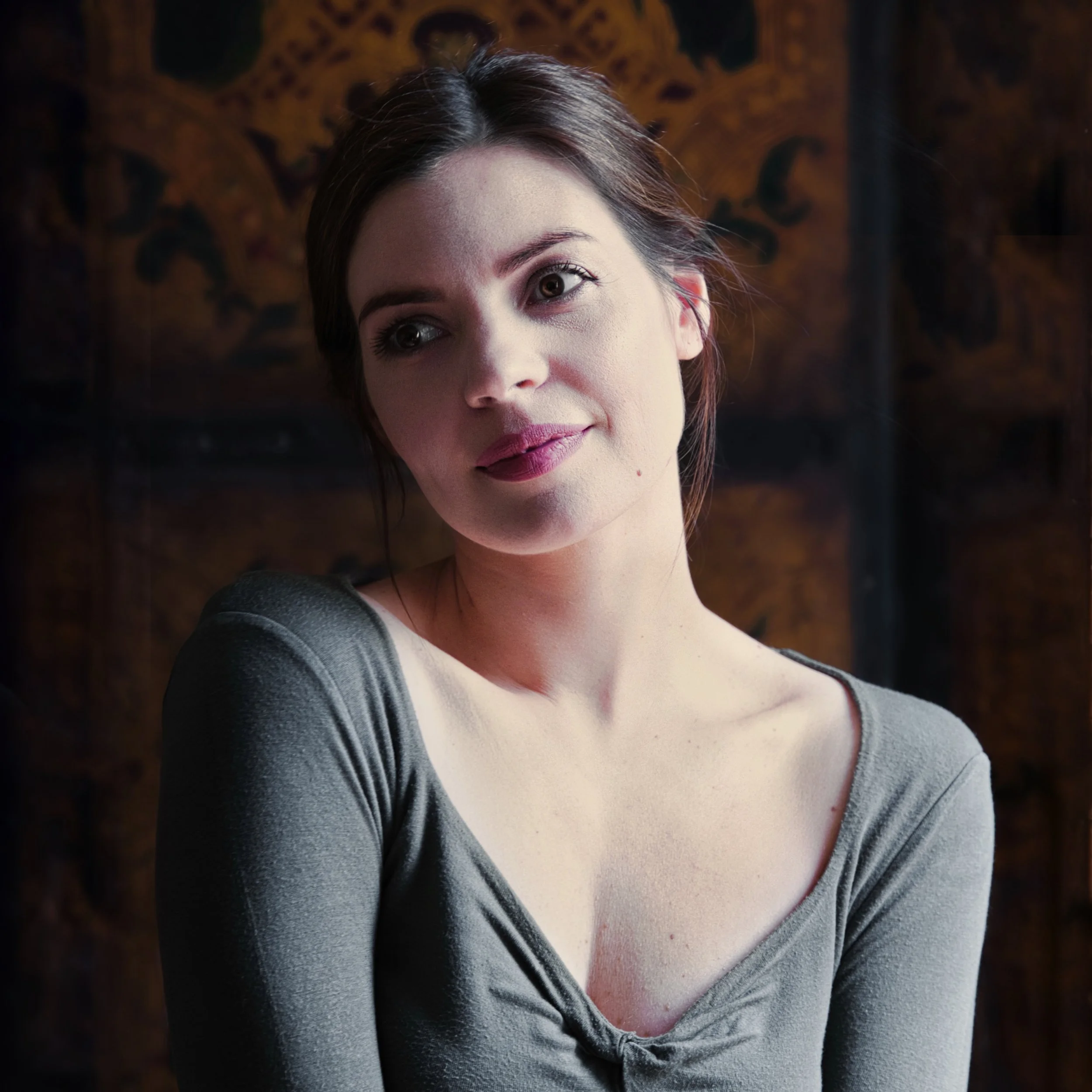 A woman with dark hair and a grey top looks towards the camera in a softly lit portrait, with a warm, dark background.