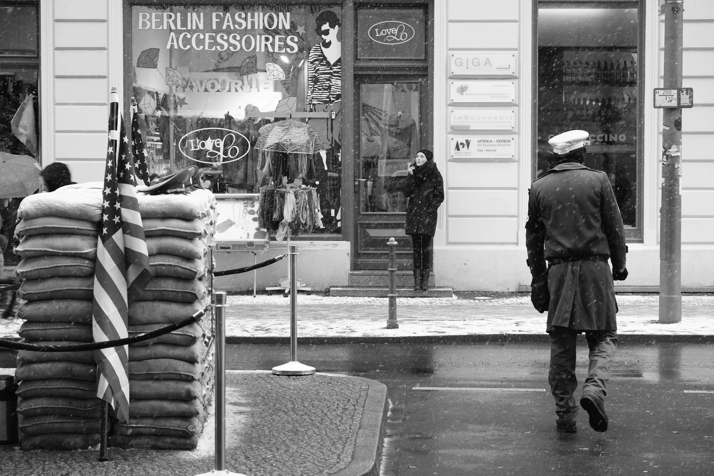 Uniformed guard walking through falling snow in Berlin while a woman stands in the doorway of a fashion accessories shop across the street