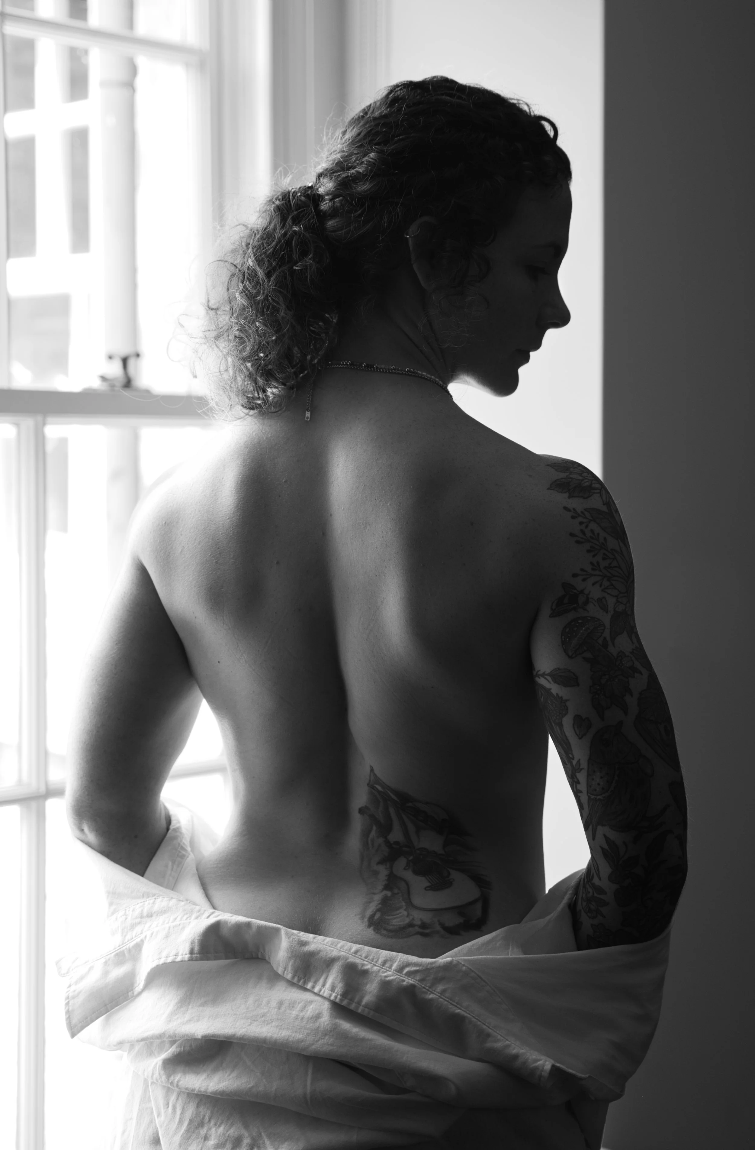 Black and white back study of a woman by a window, photographed with natural light during a boudoir session.
