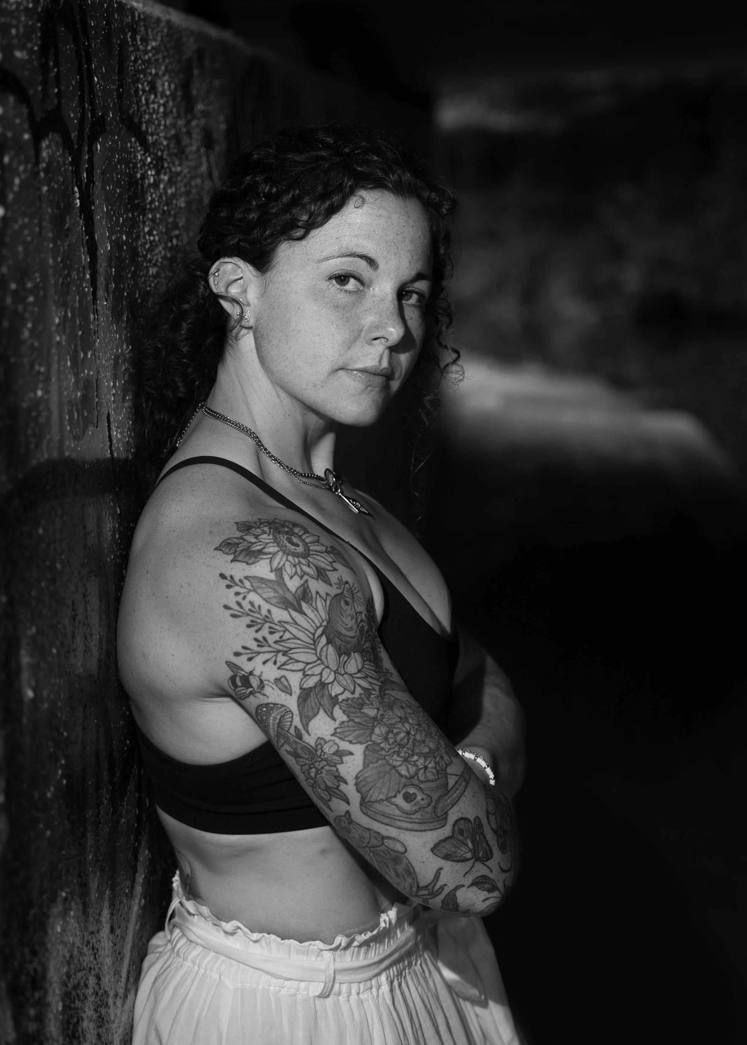 Black and white portrait of a tattooed woman leaning against a graffiti-covered underpass wall and looking towards the camera.