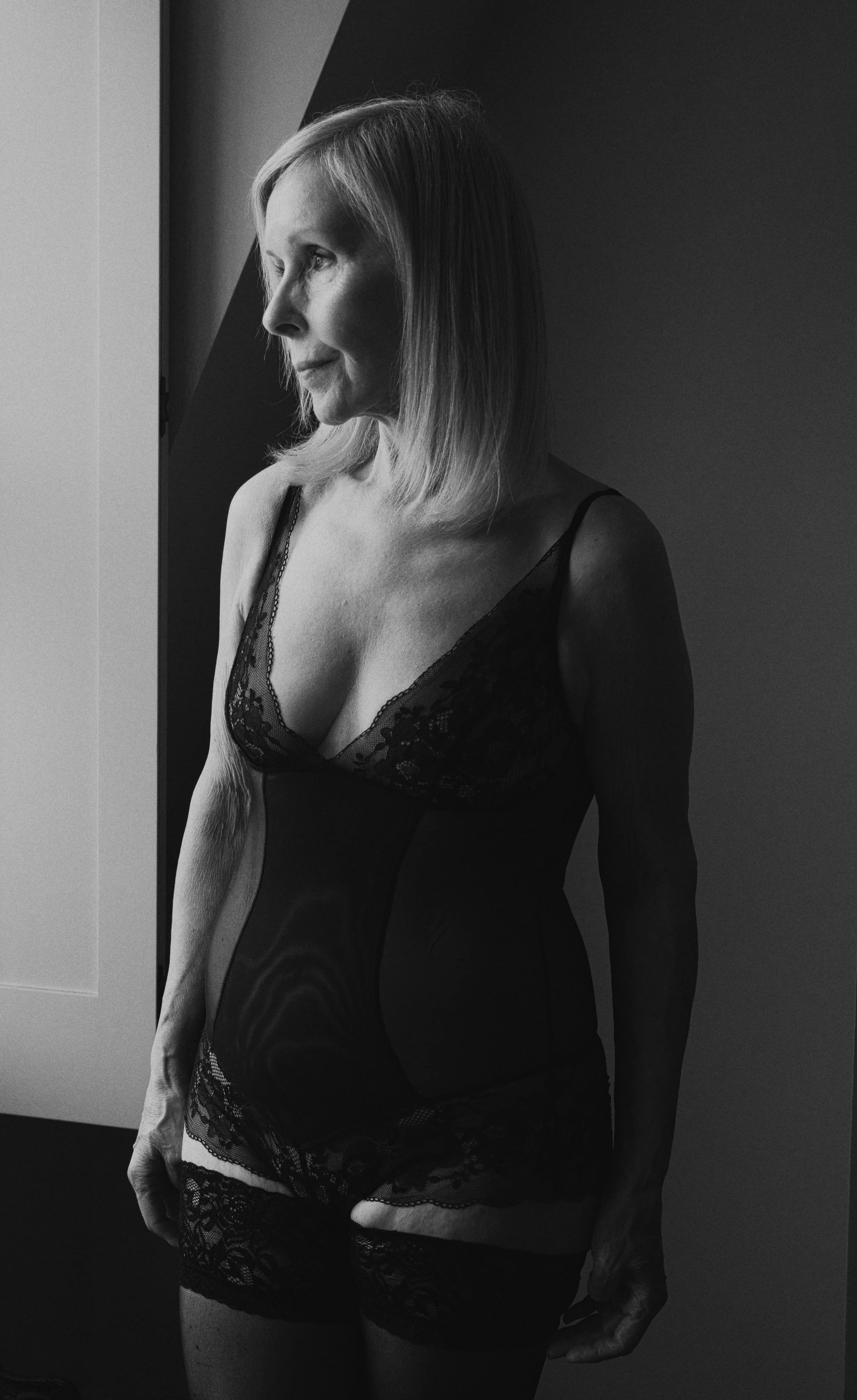 Black and white boudoir portrait of a mature blonde woman wearing black lace lingerie and stockings, standing beside a window and looking thoughtfully towards the light.