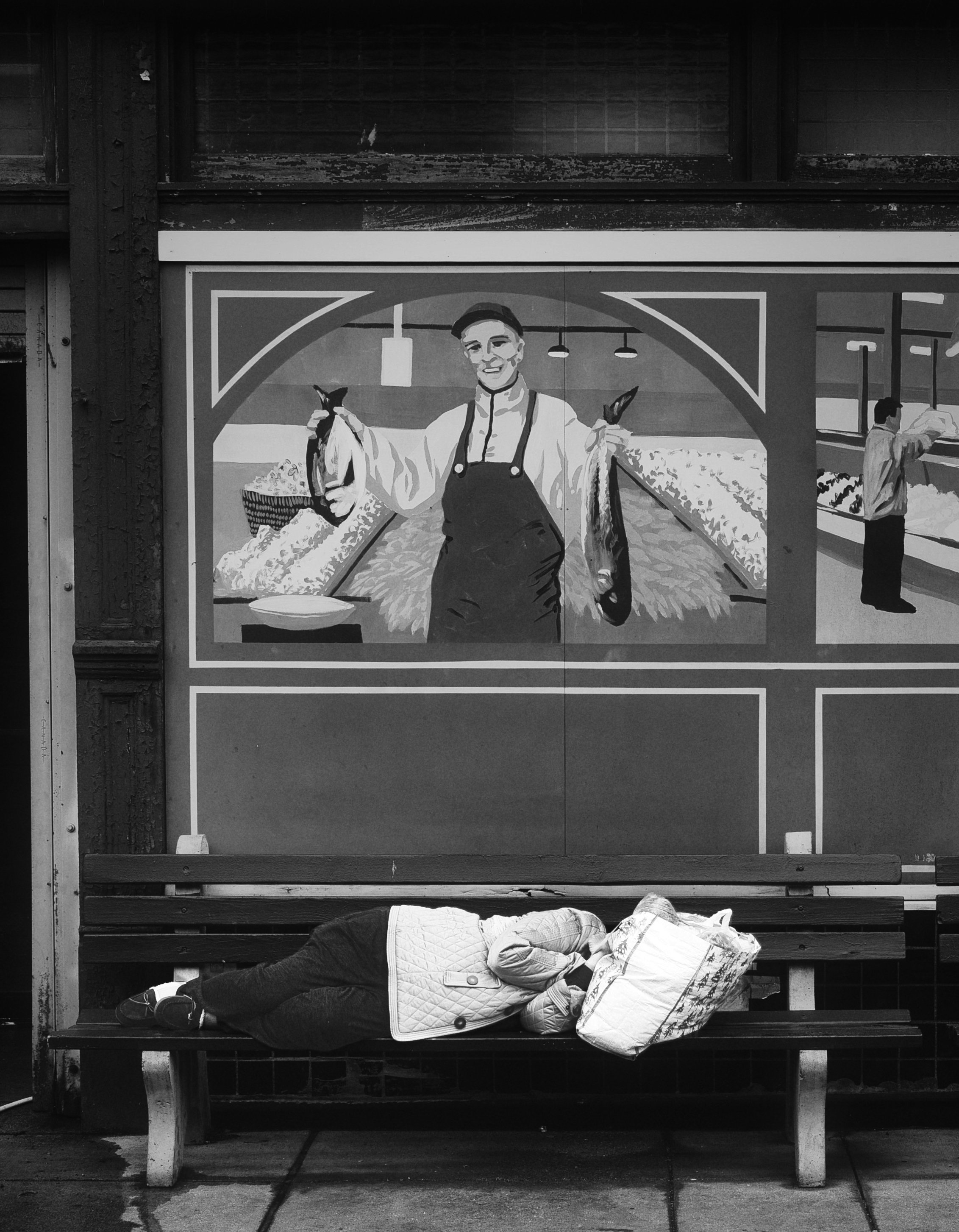 Person sleeping on a bench beneath a painted fish market mural in Cincinnati