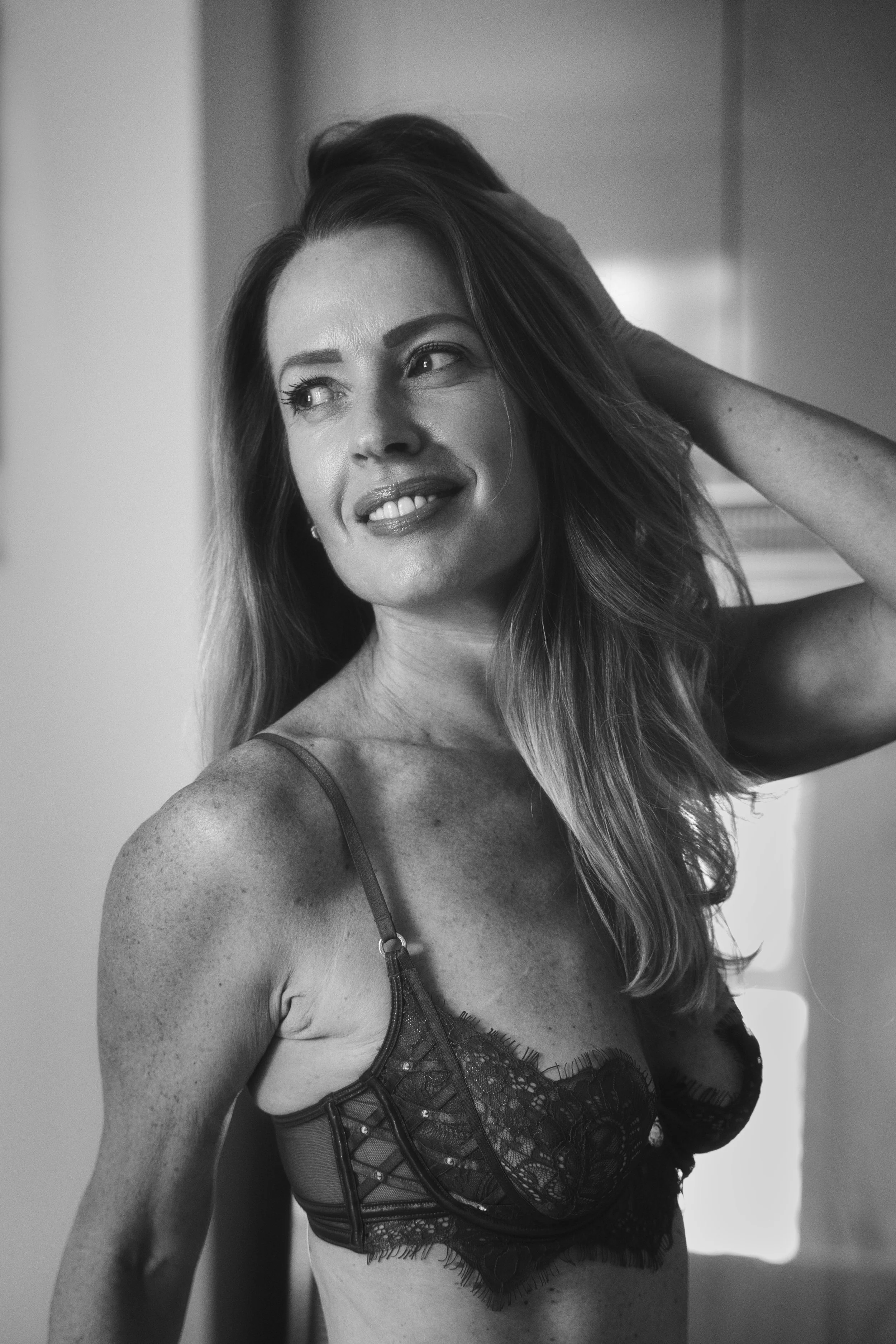 Black and white boudoir portrait of a smiling woman in lace lingerie, looking to the side with one hand resting in her hair.