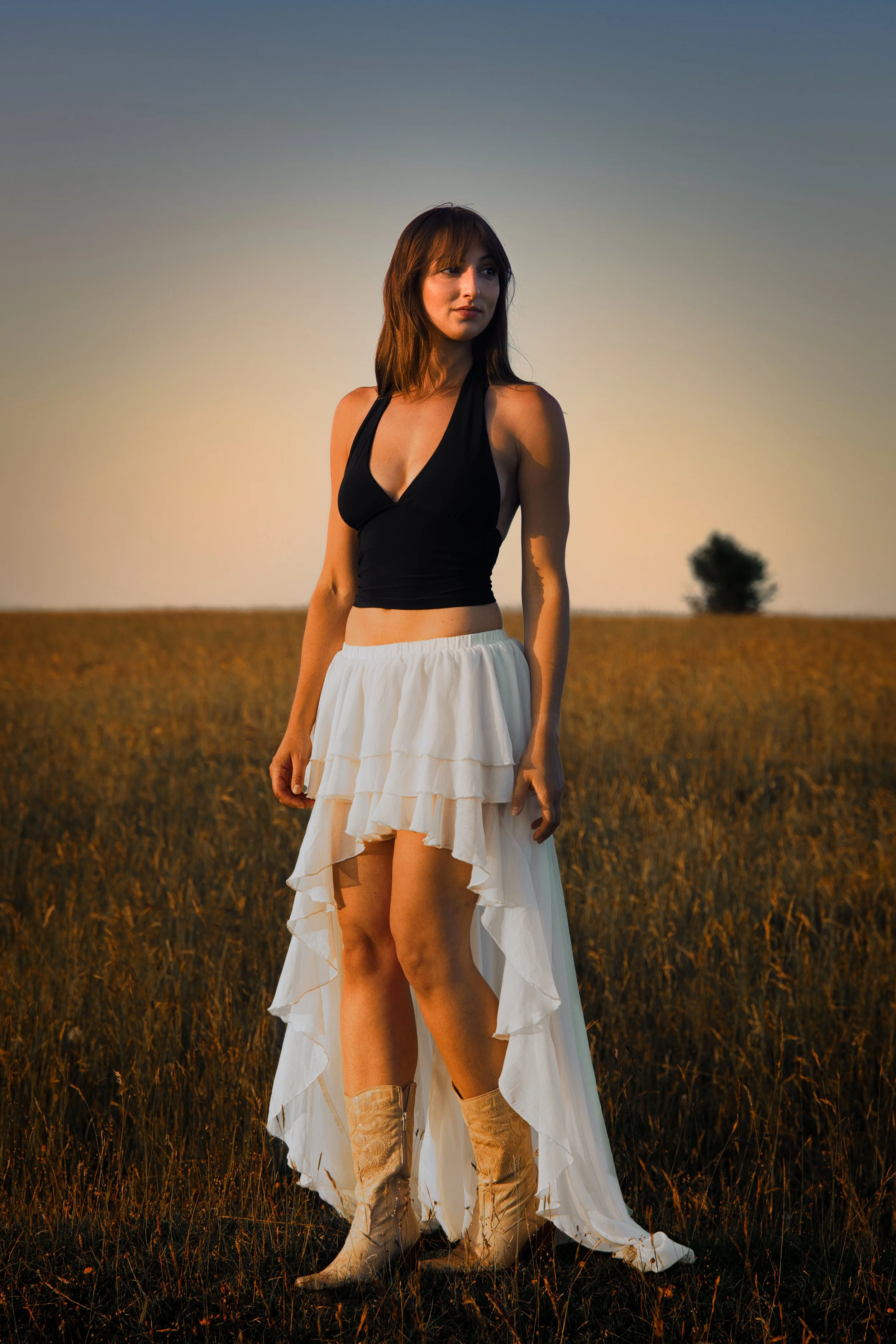Elegant full-length portrait of a woman standing in a meadow during golden hour, illuminated by warm evening sunlight against a clear sky.