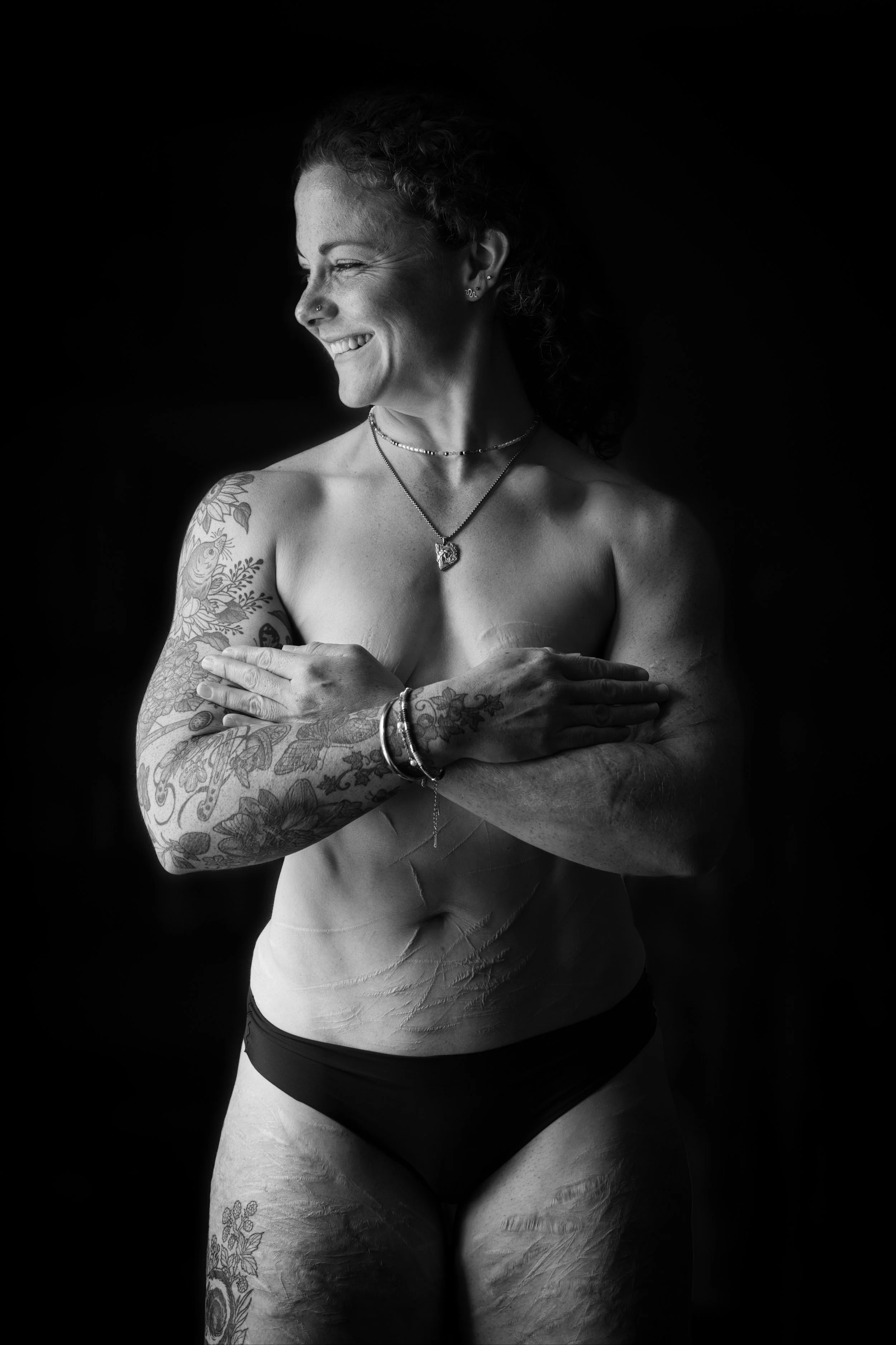 Black and white artistic nude portrait of a smiling woman with tattoos and visible scars against a dark background.