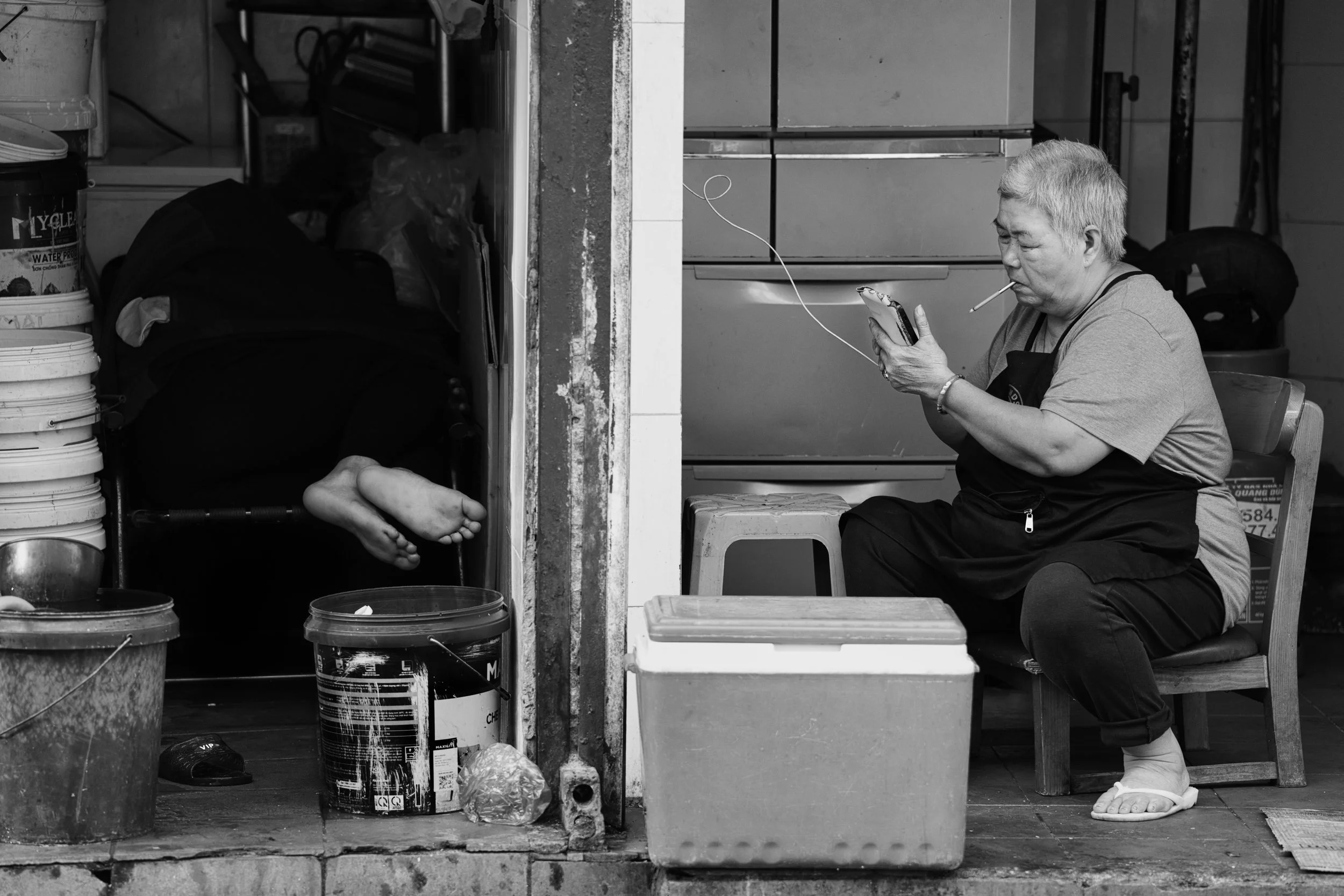 Black and white street photograph of a woman smoking while looking at her phone beside an open doorway in Hanoi