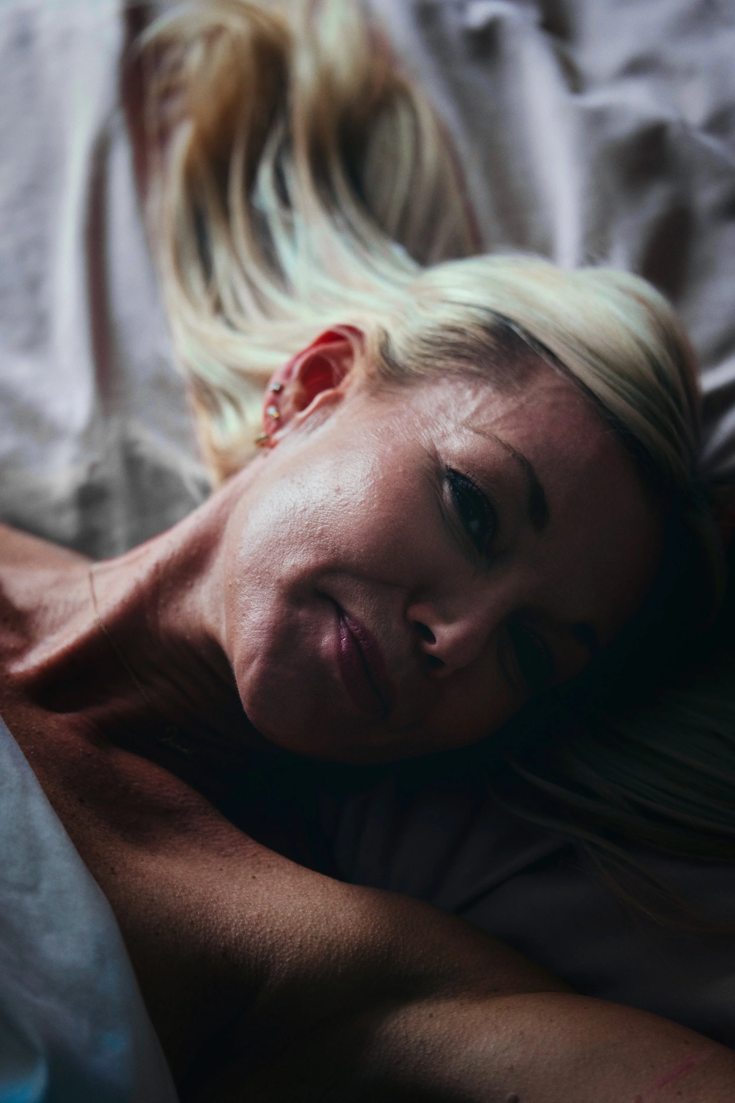 Intimate close-up boudoir portrait of a blonde woman lying in bed, softly lit with a relaxed gaze toward the camera.