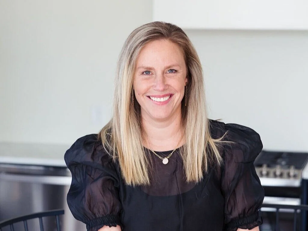 A woman with blonde hair, wearing a black puff-sleeve dress and a gold necklace, smiling in a kitchen.