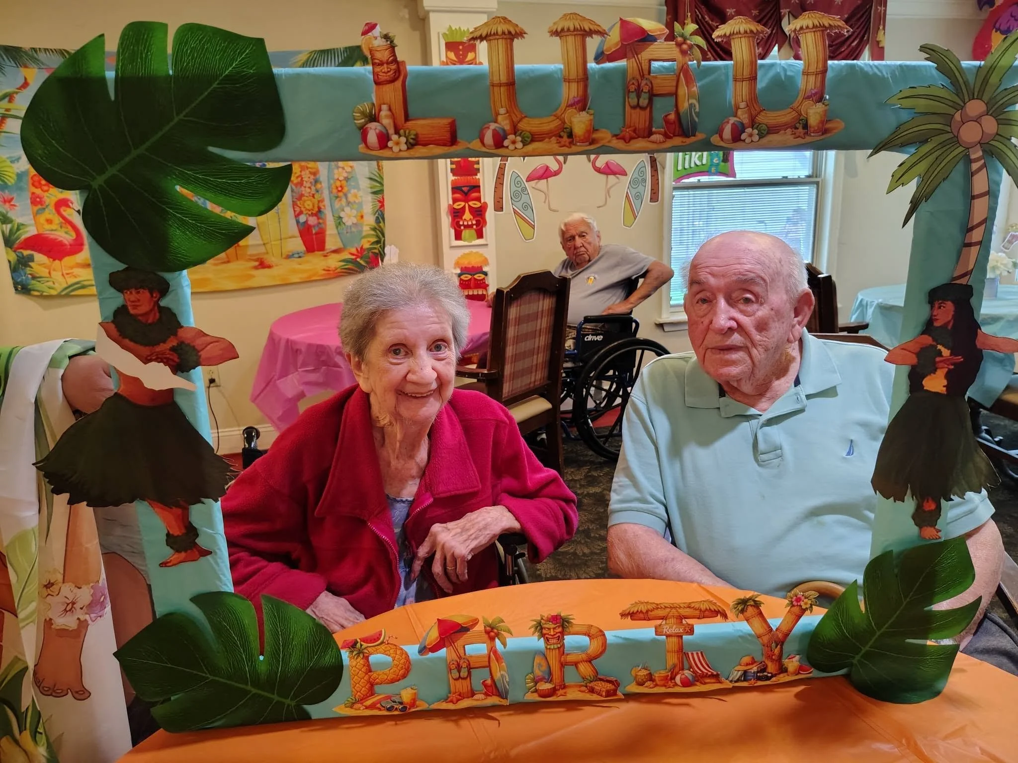 A tropical-themed party decoration with a banner that spells out "Aloha" in large wooden letters, adorned with miniature umbrellas, beach balls, and flowers, surrounded by artificial green leaves and palm trees, with elderly people sitting at a table