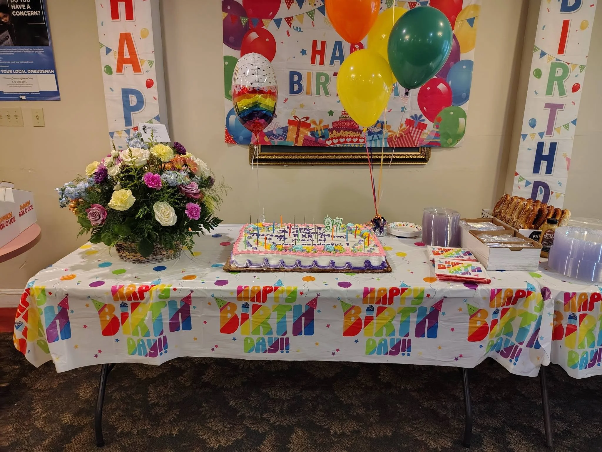 A table decorated with a colorful 'Happy Birthday' tablecloth, featuring balloons, a birthday cake, a flower bouquet, and party snacks.