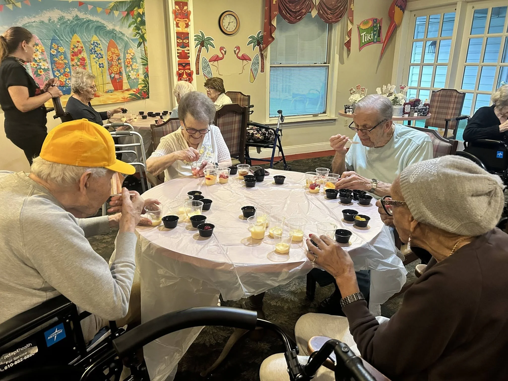 A group of elderly people sitting around a round table enjoying pudding cups, with some in wheelchairs, in a room decorated with tropical-themed wall art and banners.
