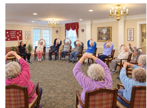 Elderly adults participating in a seated exercise class in a common room, stretching while sitting on chairs.