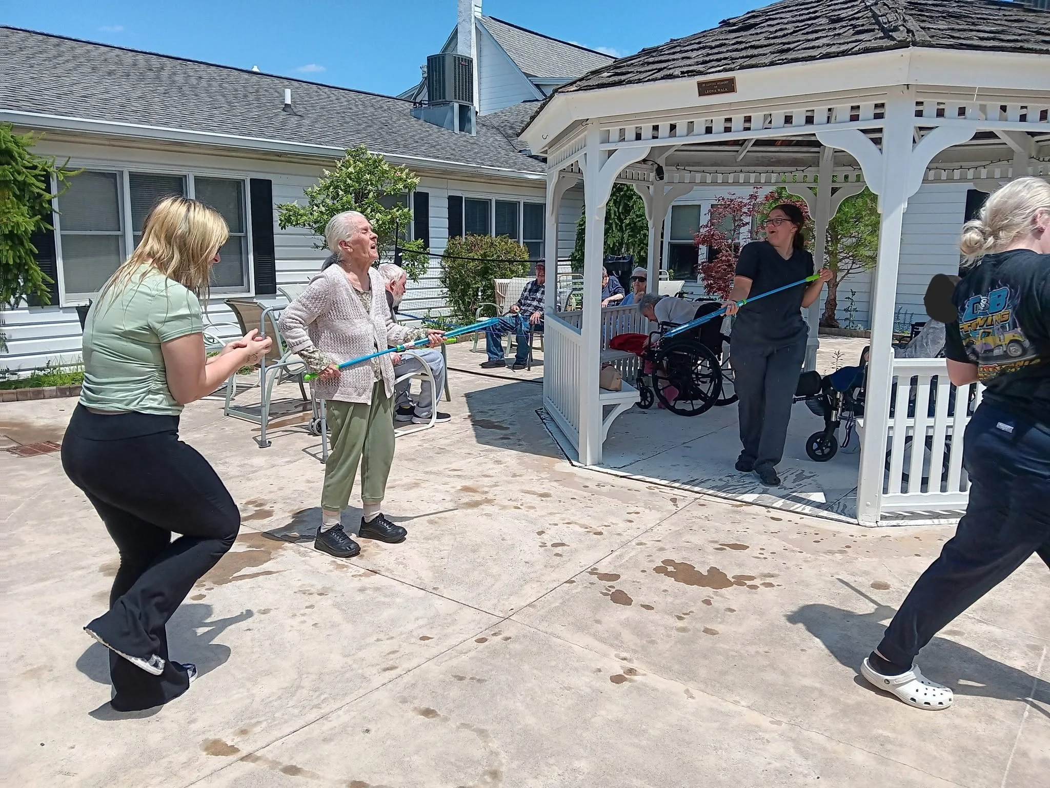 A group of people, including elderly and young individuals, are gathered outside under a gazebo, engaging in dancing and socializing on a sunny day.