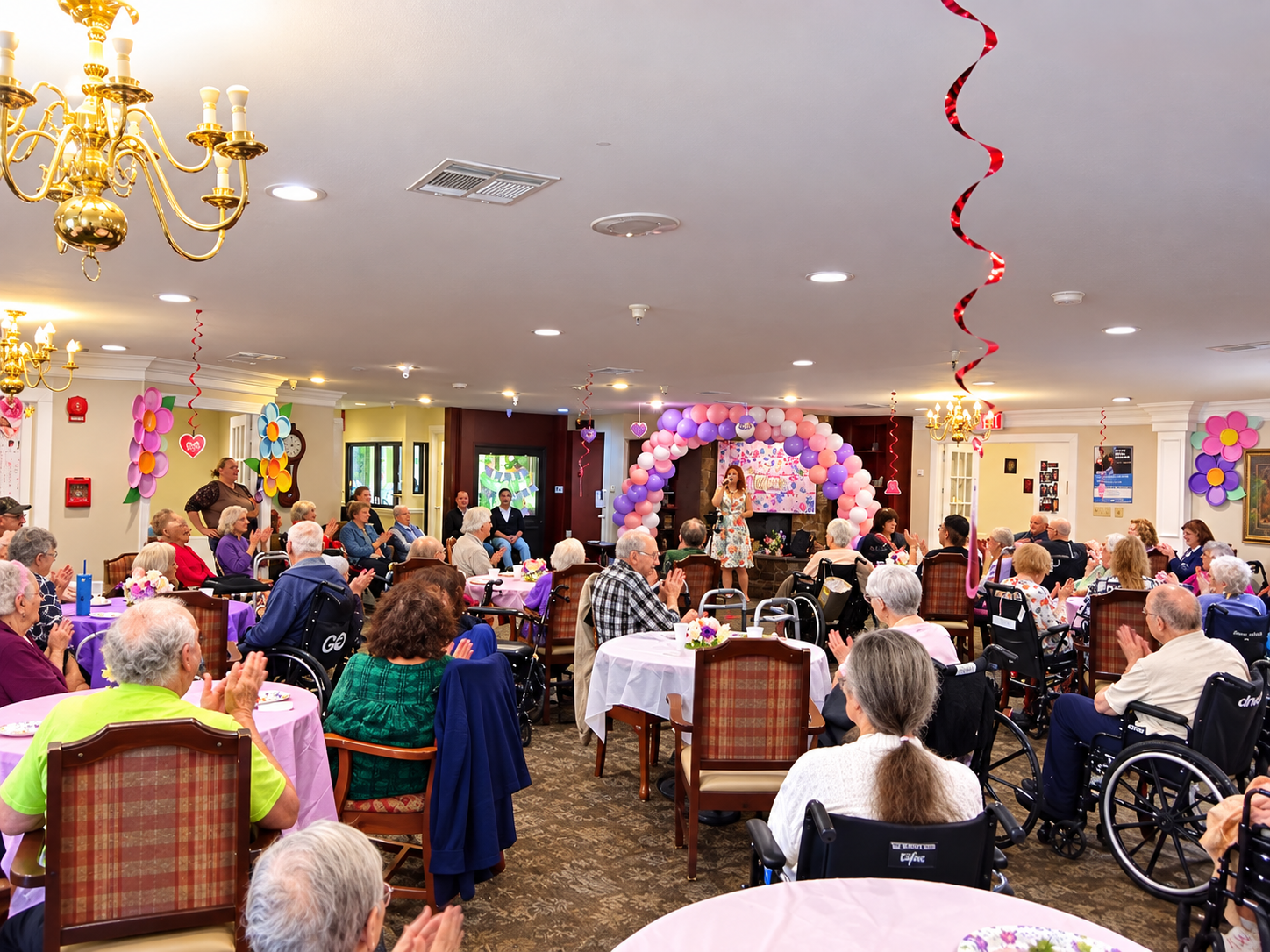 A decorated room filled with elderly people sitting at tables, clapping, and enjoying a live performance by a woman on stage. The room features balloons, streamers, and floral decorations, creating a festive atmosphere.