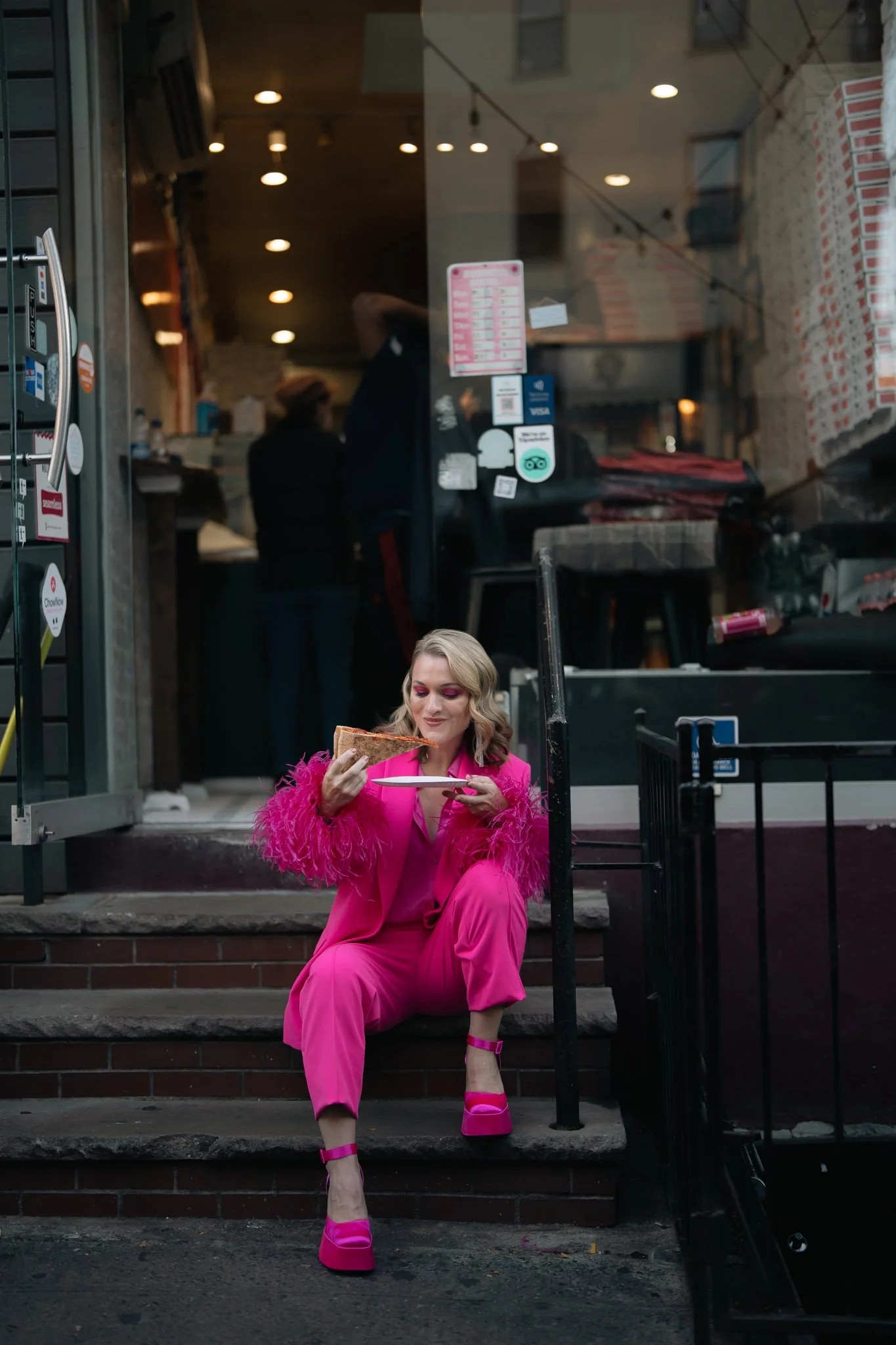 A woman dressed in a bright pink suit with feathered cuffs and matching pink high heels, sitting on the steps outside a restaurant or cafe, eating a slice of pizza.