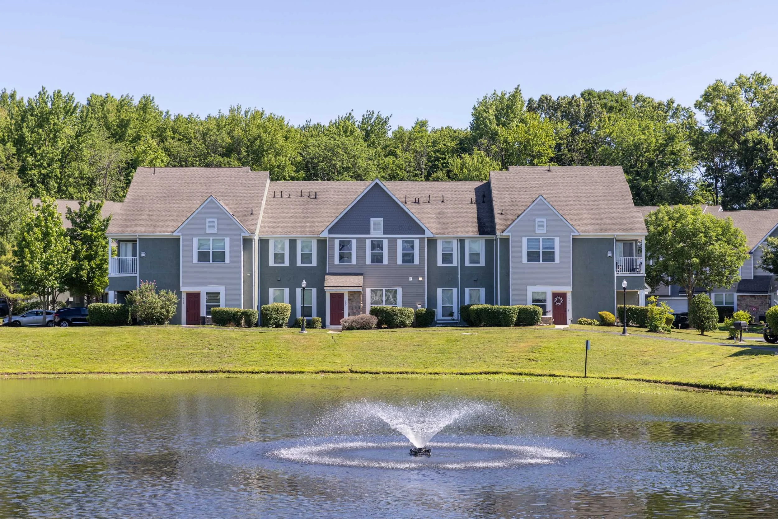 A row of apartments with gray siding, red doors, and a small front porch, situated behind a pond with a fountain, green lawn, bushes, and trees in the background on a sunny day.