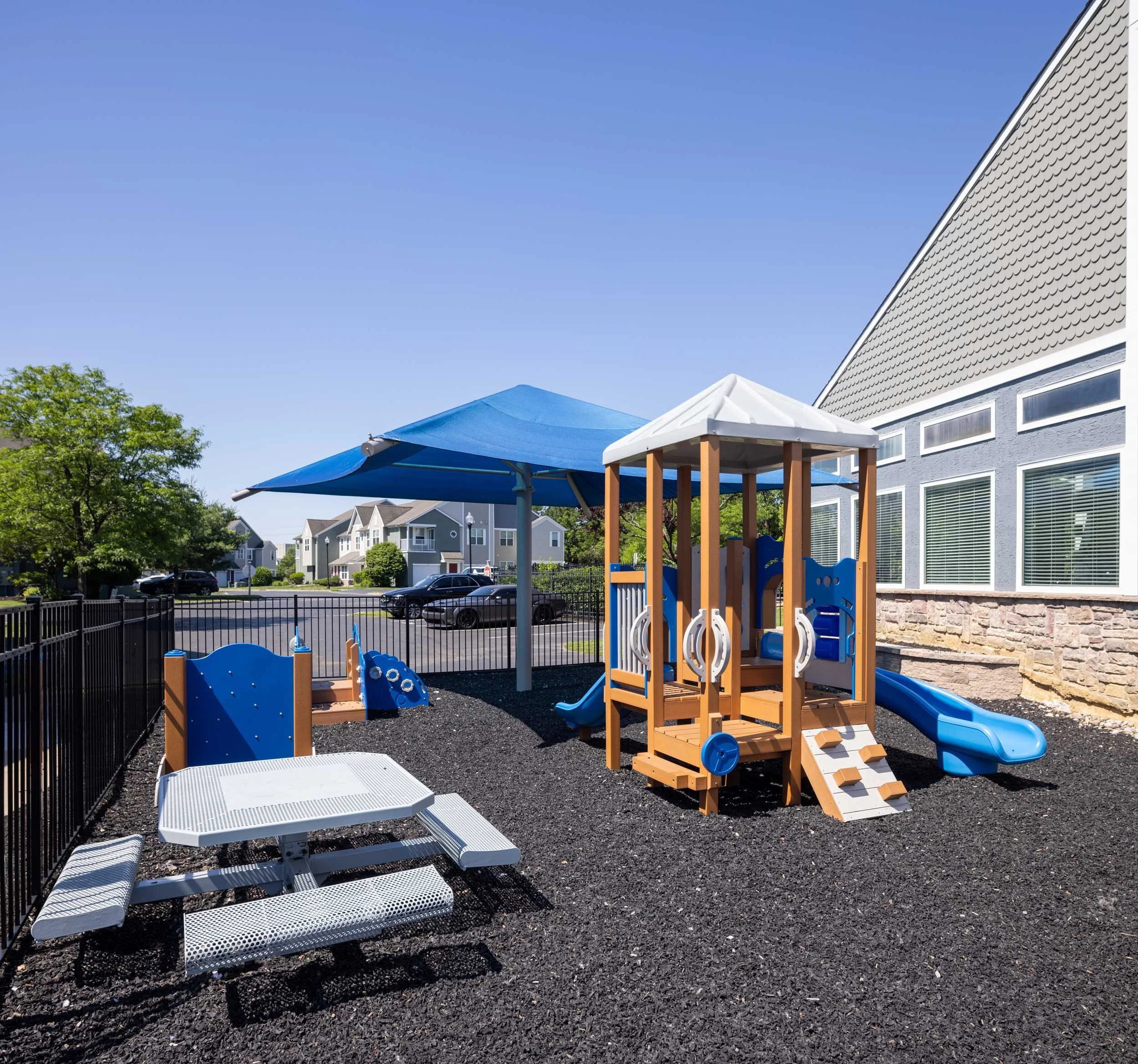 Children's playground with a blue shade canopy, white picnic table, and a play structure with a slide, surrounded by a black fence, with apartment buildings and green trees in the background on a sunny day.