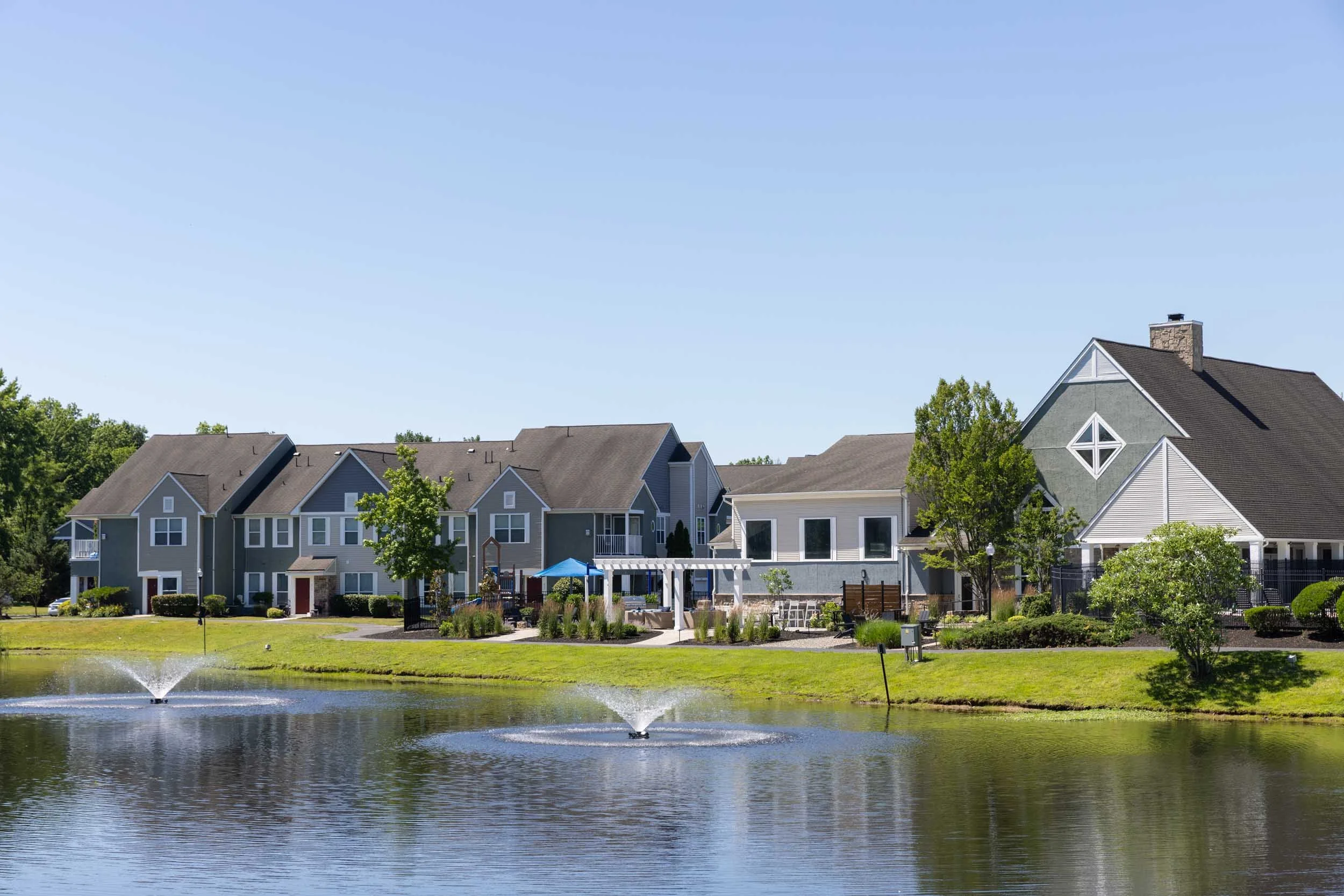 A neighborhood with multiple gray and white apartments with sloped roofs, surrounded by trees, and featuring a small pond with fountains in the foreground under a clear blue sky.