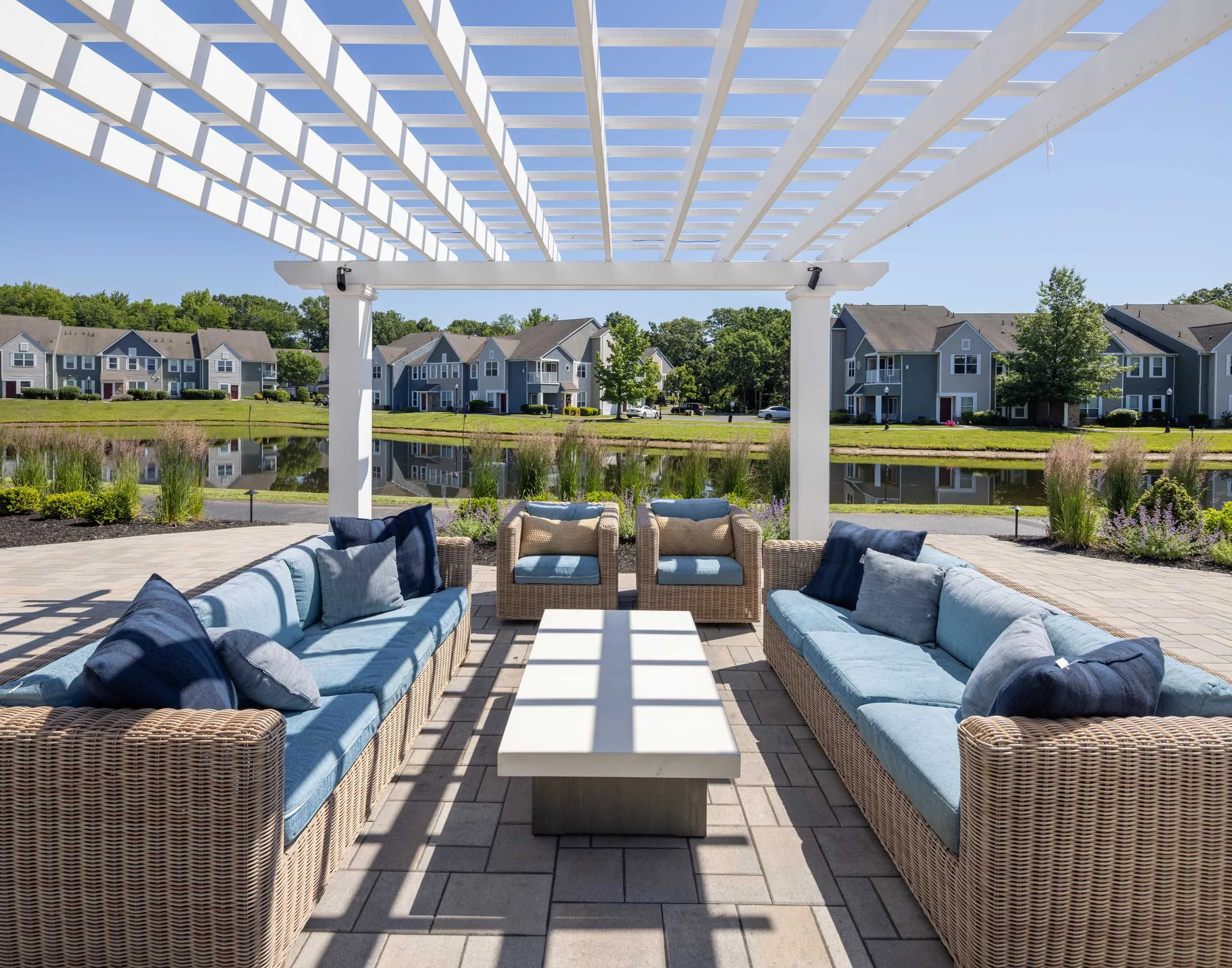 Outdoor seating area with wicker sofas and blue cushions, shaded by a white pergola, overlooking a pond with surrounded by houses and greenery on a sunny day.