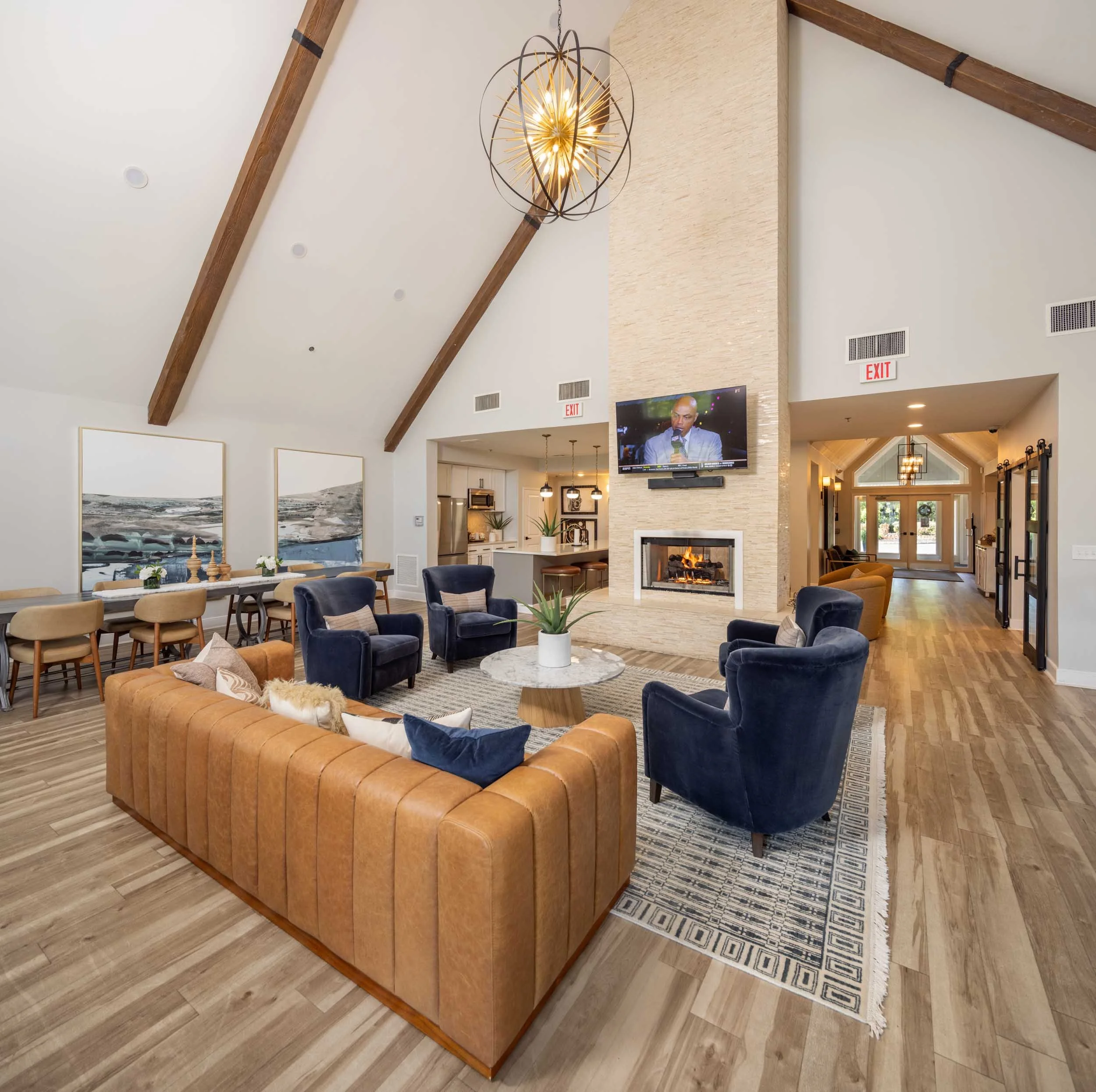 Interior view of a club room with a vaulted ceiling, wooden beams, a modern chandelier, a fireplace, a dining table, and comfortable chairs.