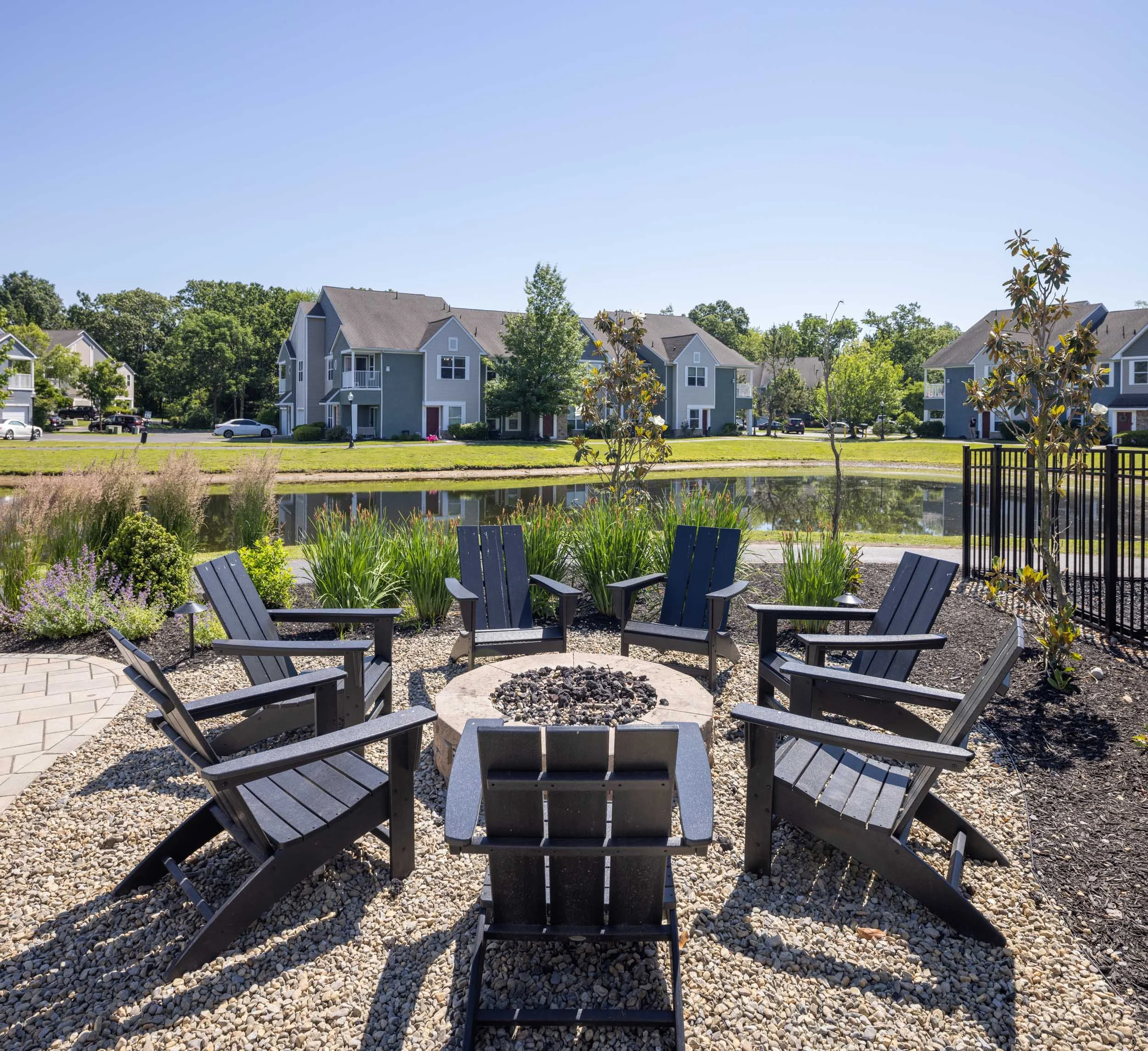 Outdoor seating area with Adirondack chairs arranged around a fire pit on a gravel surface, overlooking a pond with a landscaped yard and houses in the background on a sunny day.
