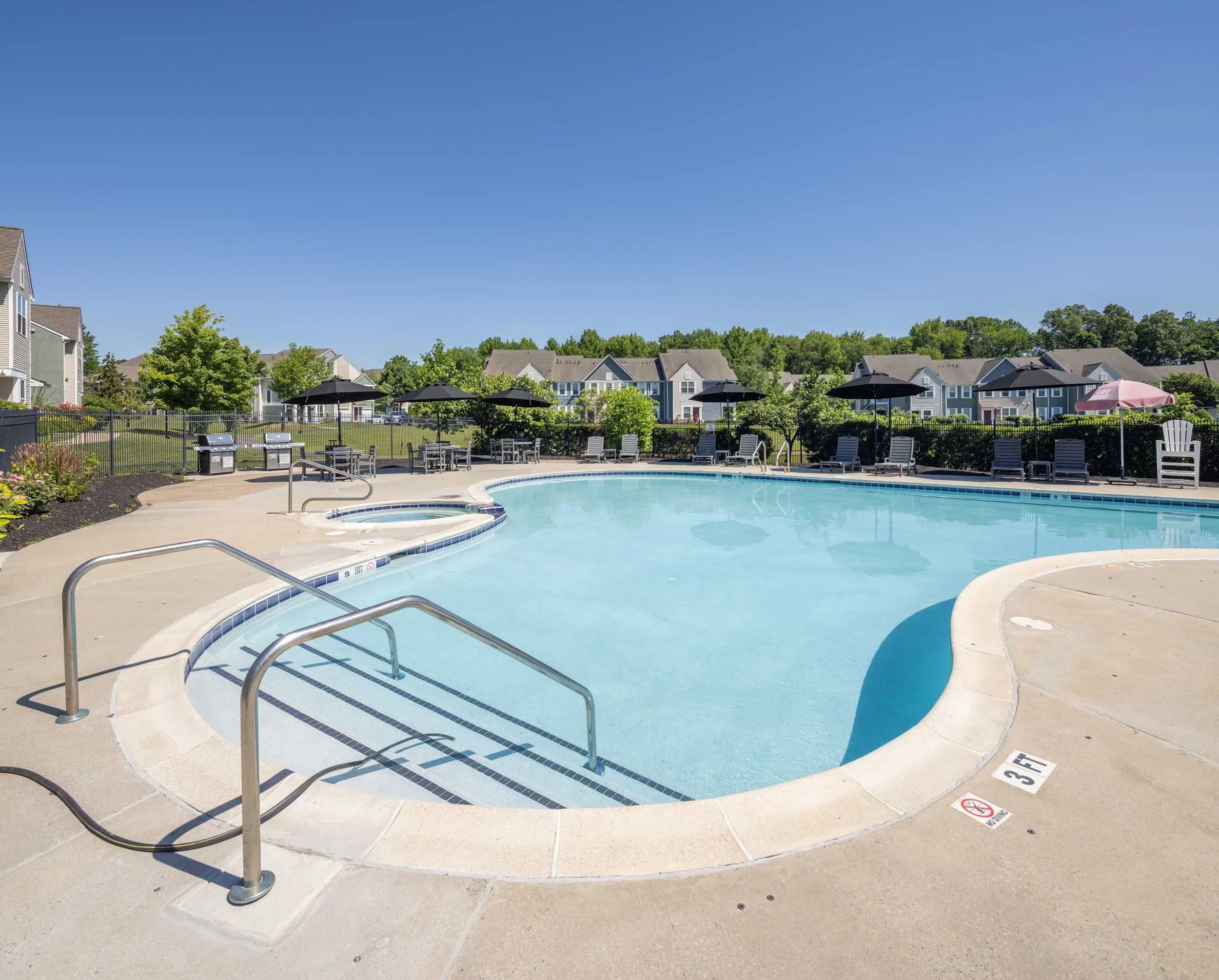 Community swimming pool with chairs, umbrellas, and a fence, surrounded by apartment buildings and green trees, under a clear blue sky.