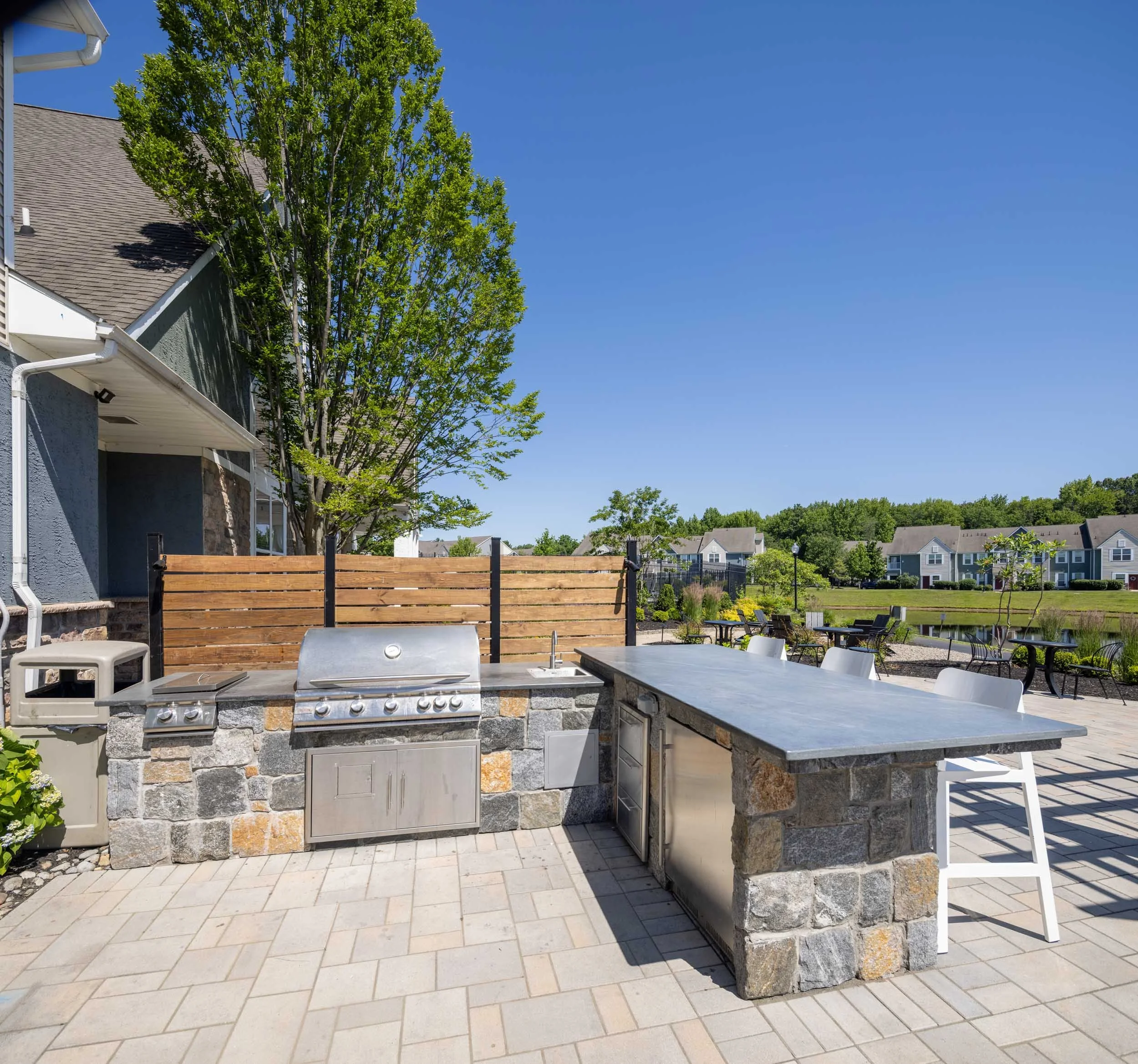 An outdoor kitchen with a stone countertop, stainless steel grill, and a cabinet underneath, situated on a paved patio. In the background, there is a wooden fence, patio chairs, and a well-maintained yard with grass, trees, and townhouses.
