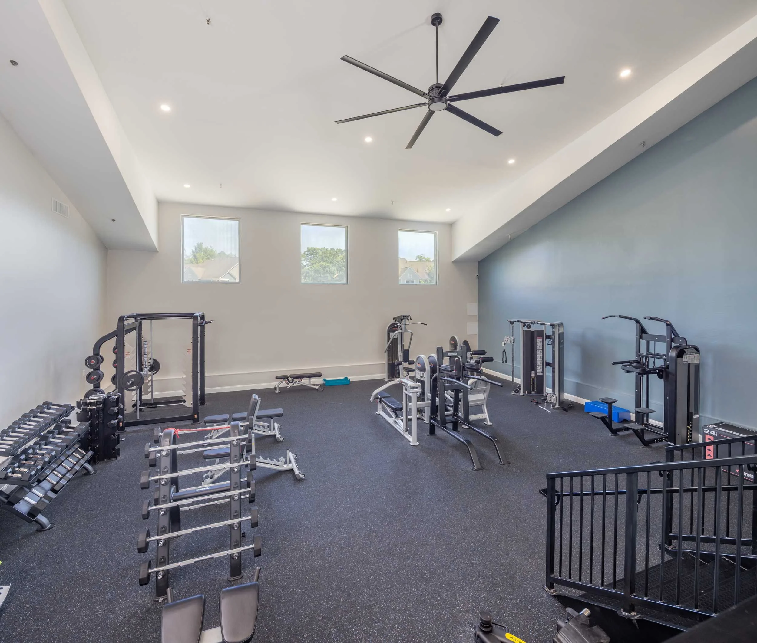 Gym with fitness equipment including dumbbells, weight benches, and cardio machines, in a bright room with large windows and a modern ceiling fan.