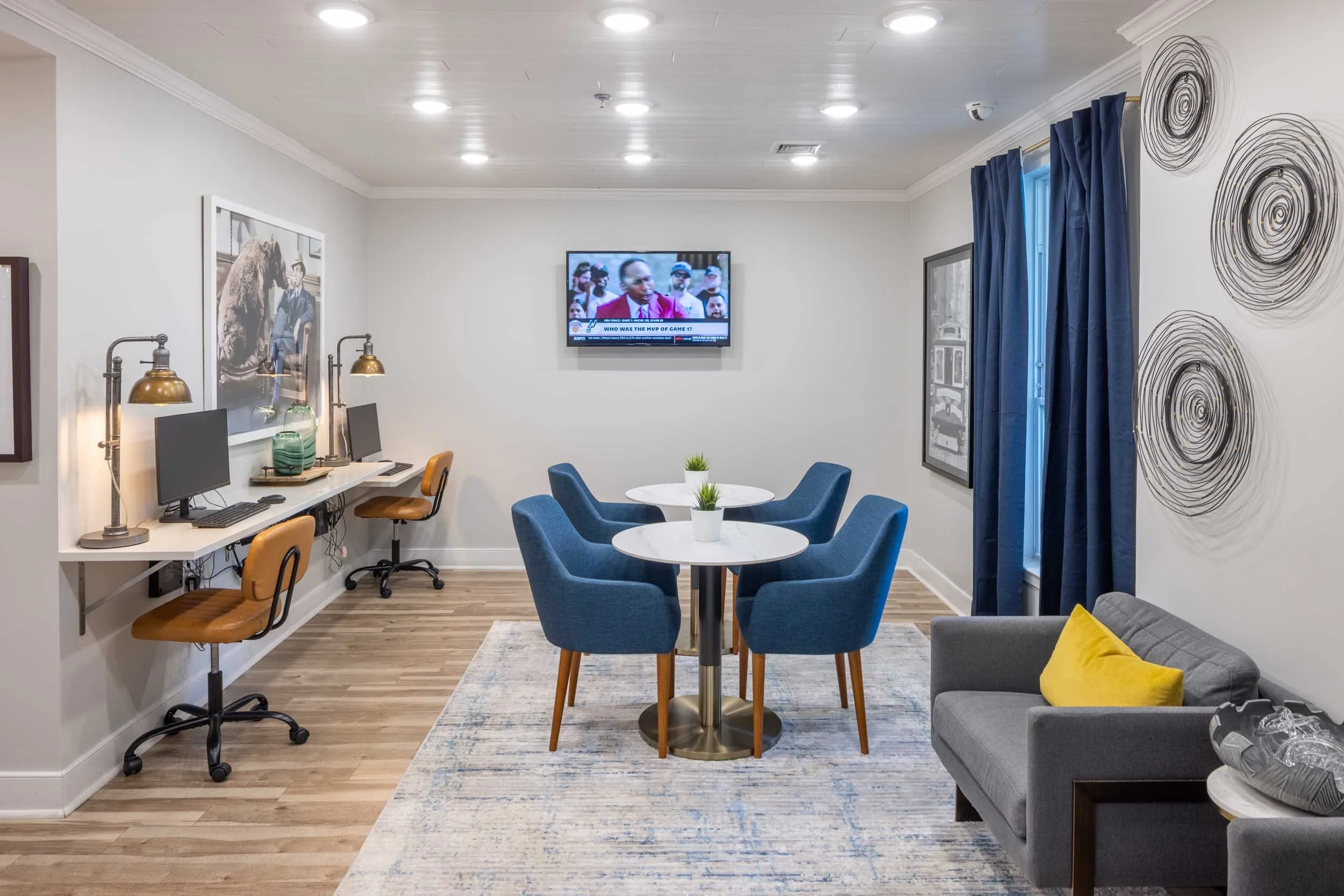 A modern indoor lounge area with a TV mounted on white wall, blue curtains on the window, round dining table with blue chairs, a corner sofa with yellow pillow, wood flooring, and decorative art pieces.